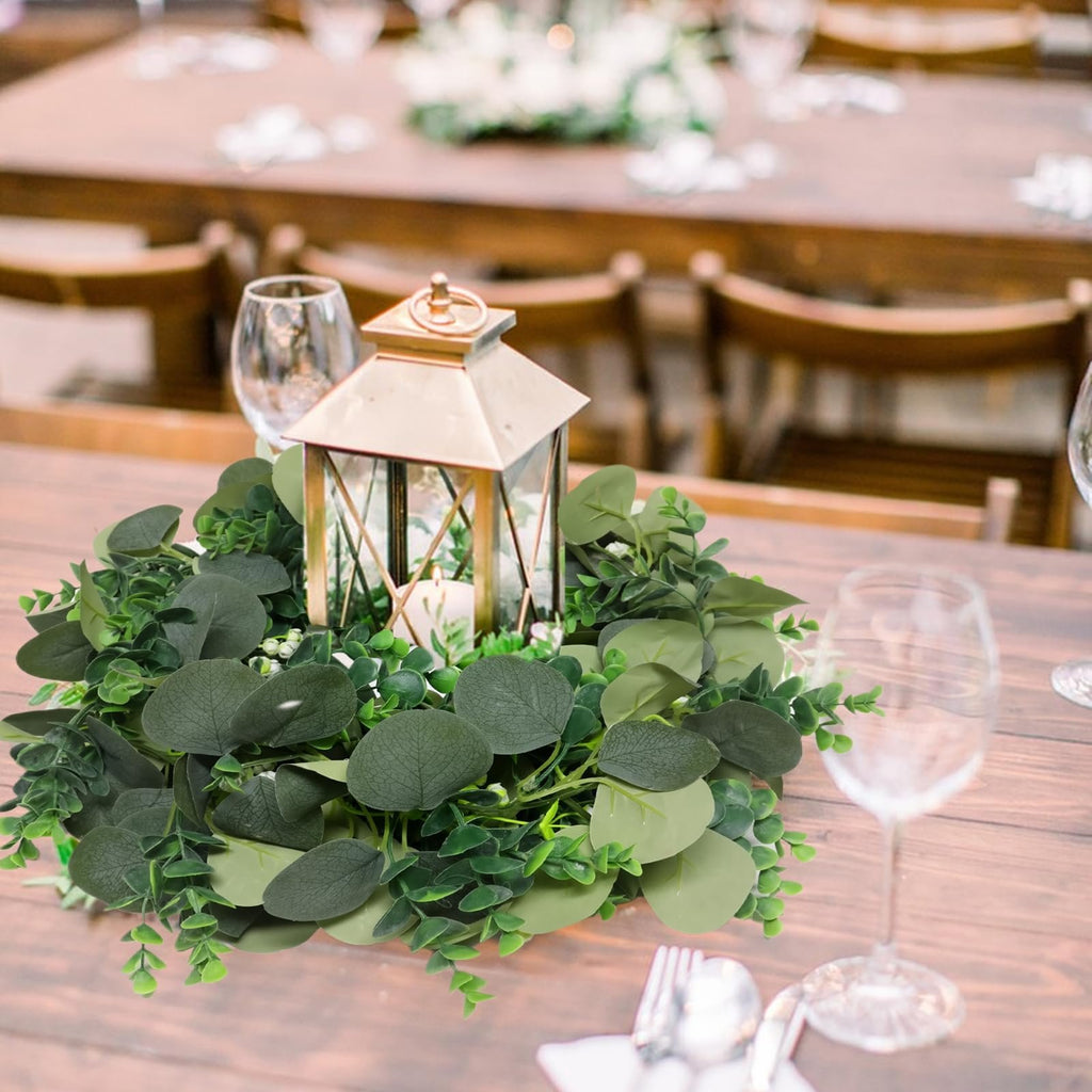 Artificial eucalyptus garland with white blooms arranged around a lantern on a wooden table with wine glasses and cutlery.