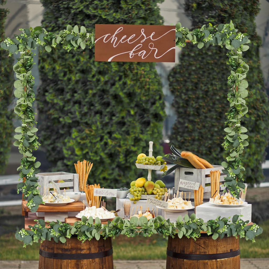 Cheese bar setup with artificial eucalyptus garland, fruits, breadsticks, and various cheeses displayed on wooden crates and barrels.
