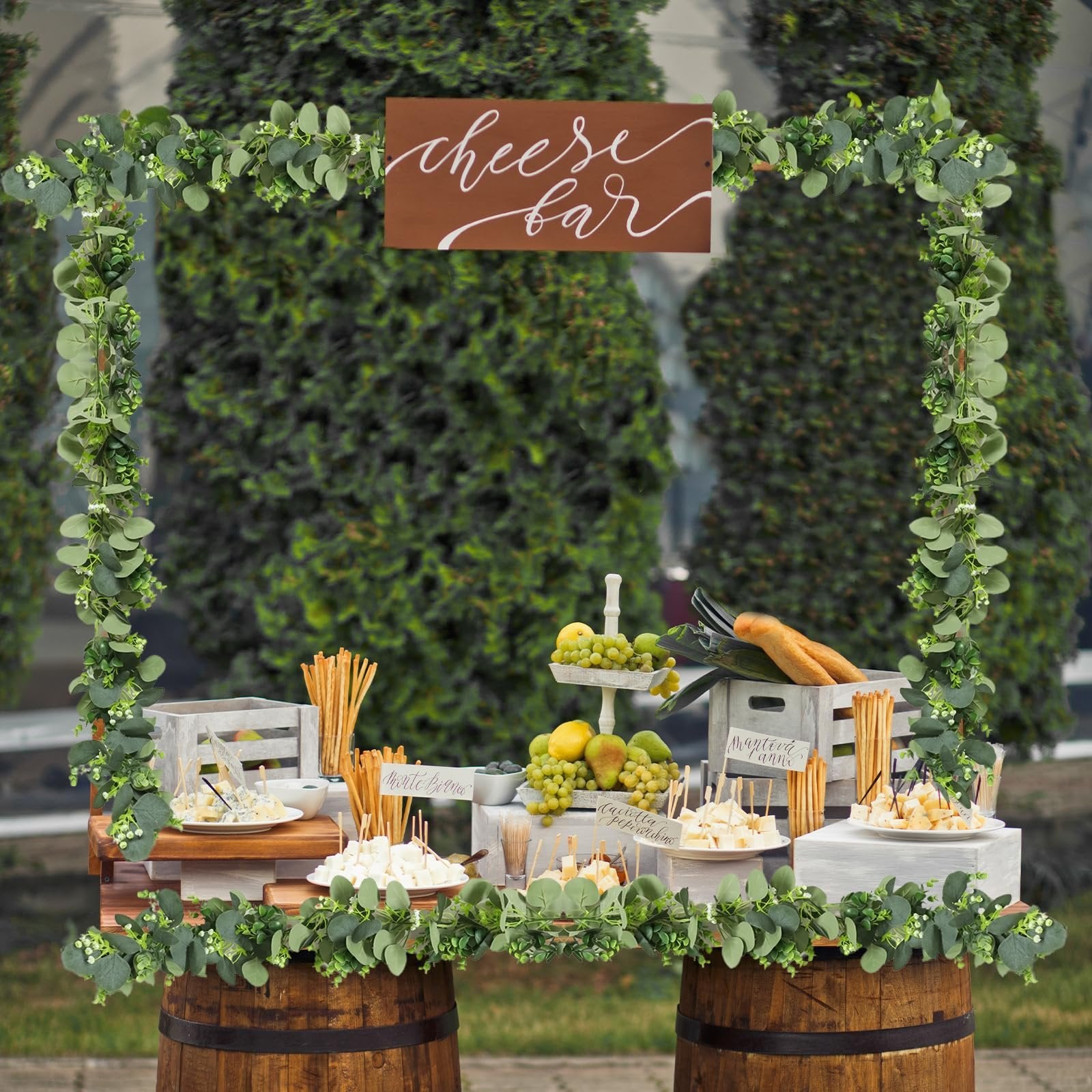 Cheese bar setup with artificial eucalyptus garland, fruits, breadsticks, and various cheeses displayed on wooden crates and barrels.