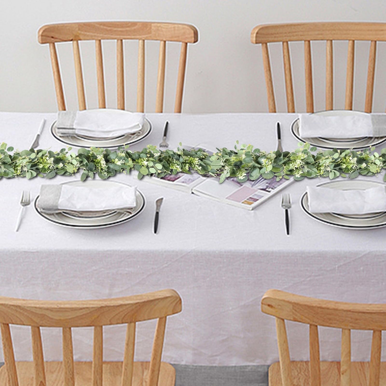 Artificial eucalyptus garland with white blooms draped across a table set for dinner with wooden chairs.