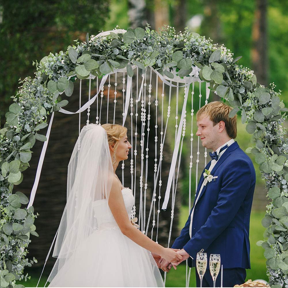 Bride and groom holding hands under a faux eucalyptus garland wedding arch decorated with white ribbons and beads.