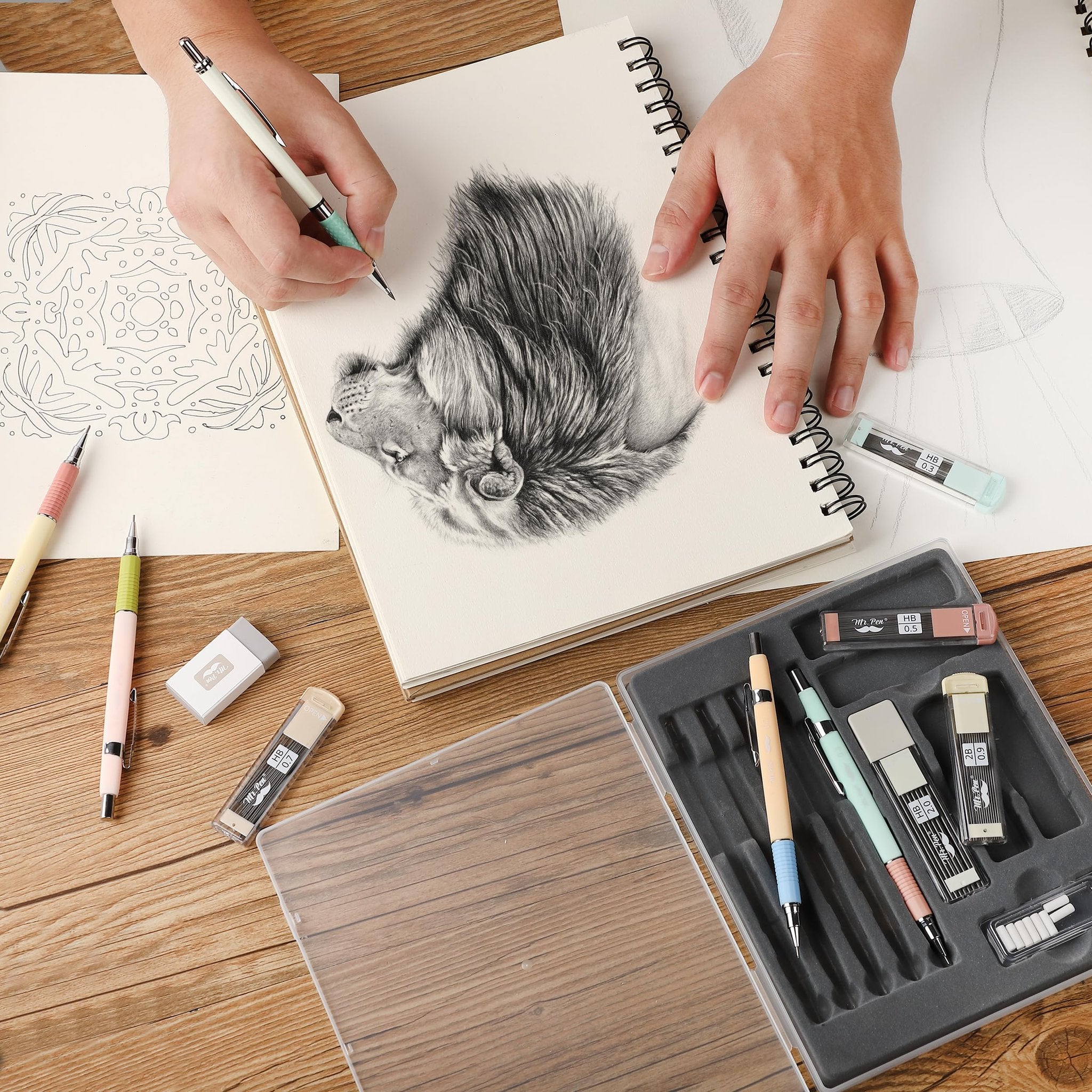 Hands drawing a lion in a sketchbook with a set of mechanical pencils, lead refills, and erasers on a wooden table.