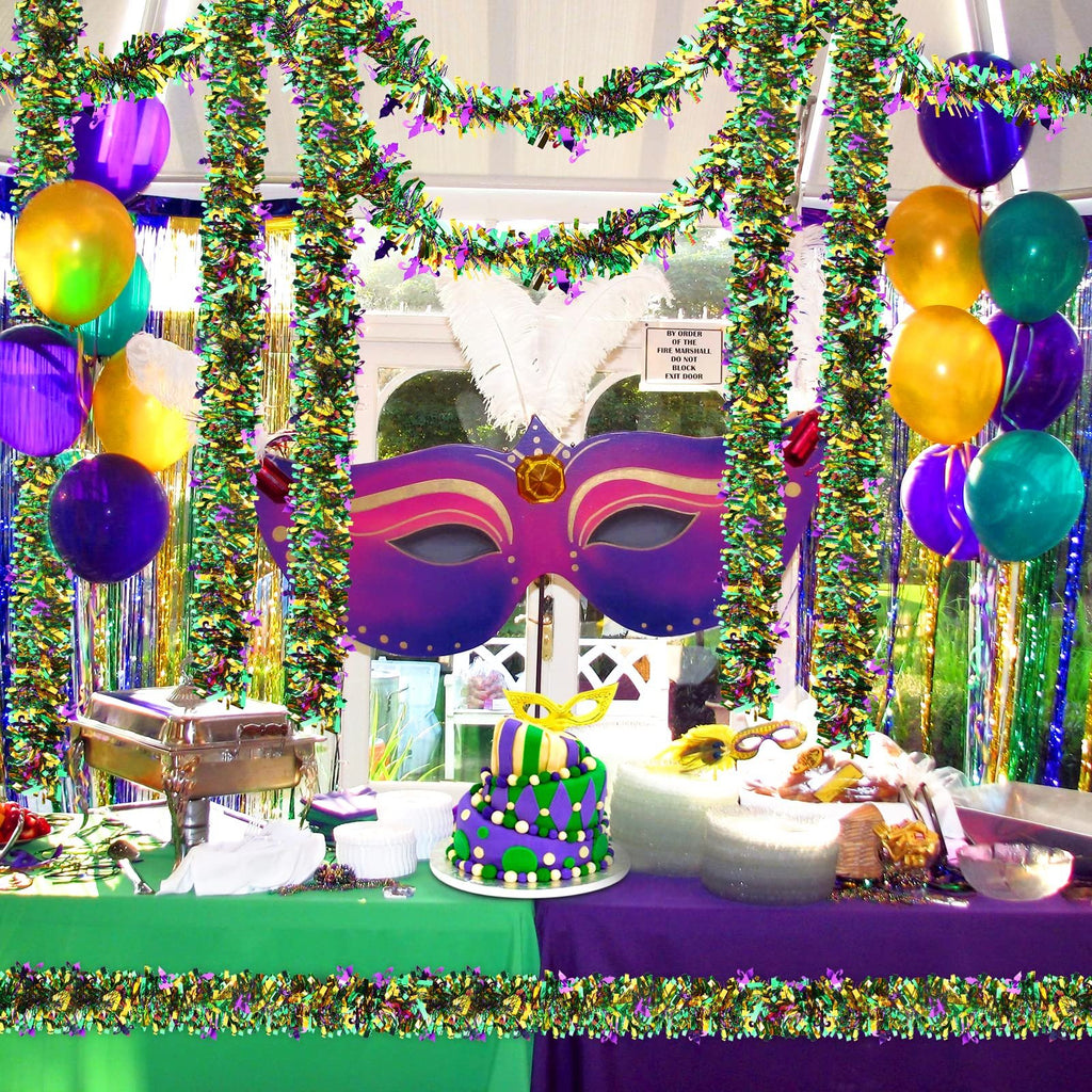 Mardi Gras party table setup with purple, green, and gold tinsel garlands, balloons, a masquerade mask, and a tiered cake.