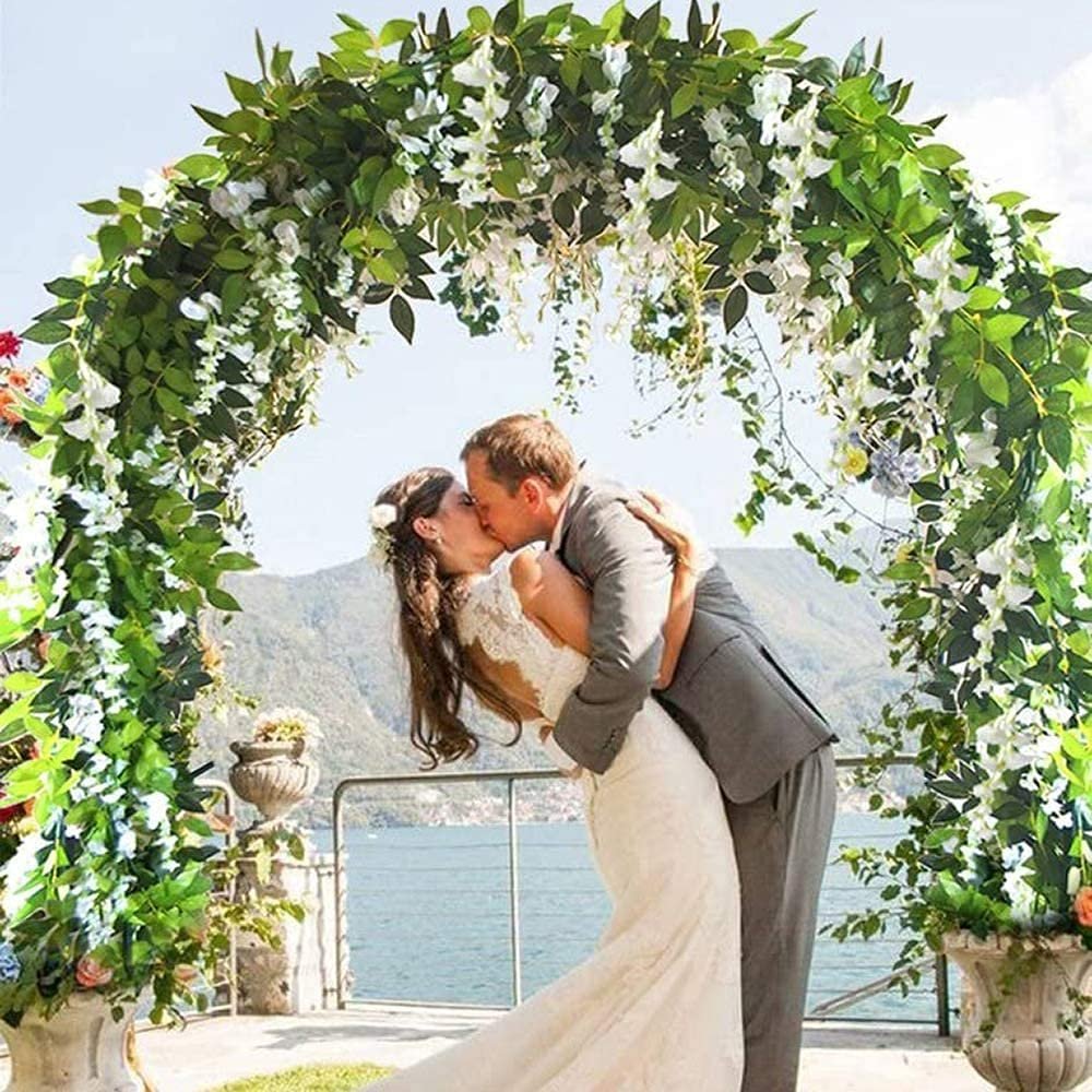 Couple kissing under a wedding arch decorated with artificial wisteria vines and greenery, overlooking a lake and mountains.