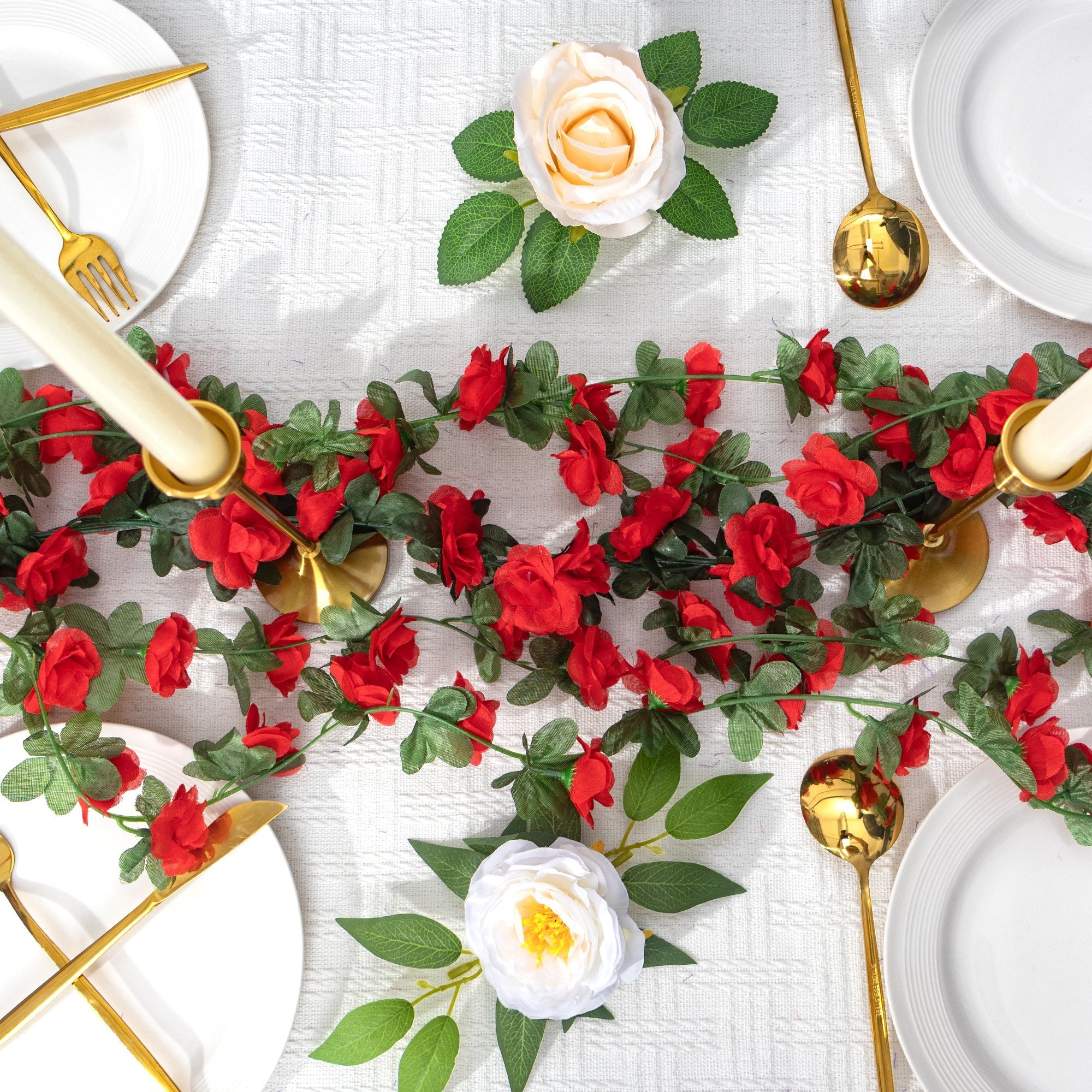 Artificial red rose garland with green leaves draped across a table setting with white plates and gold cutlery