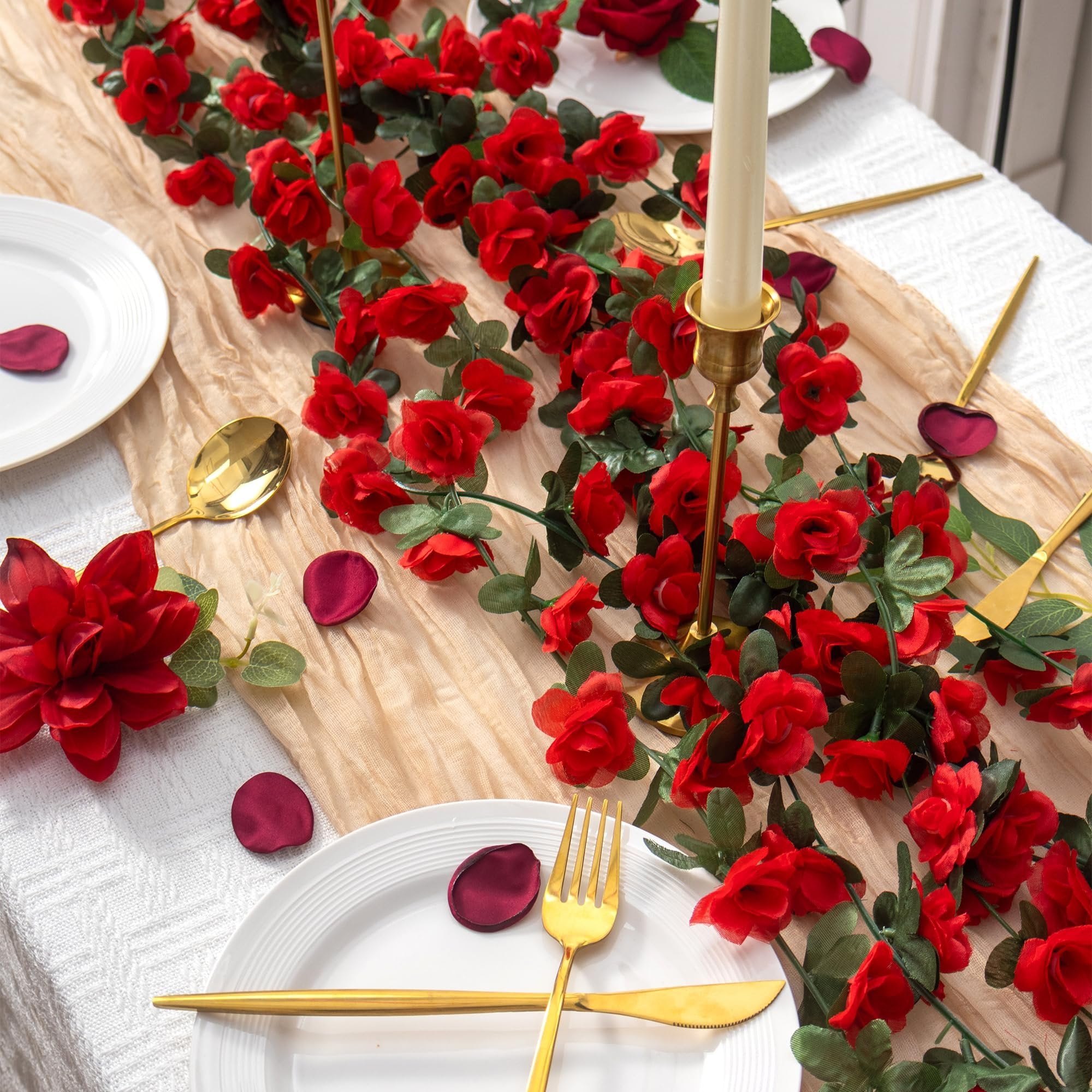 Artificial red rose garland with green leaves draped across a table setting with plates, gold cutlery, and a candle.