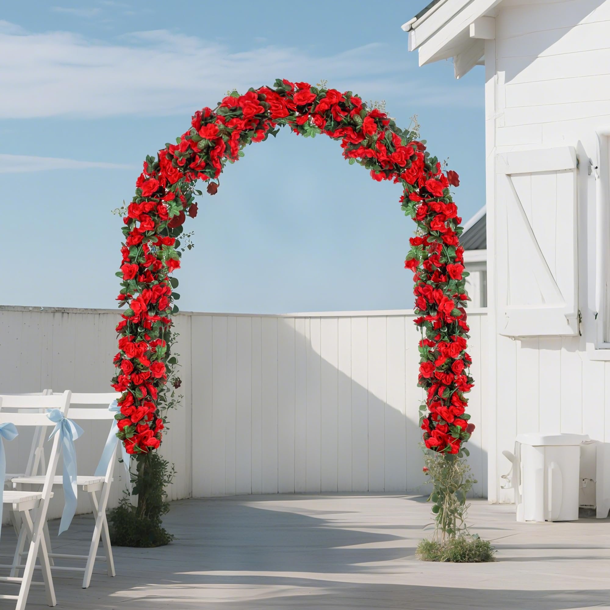 Artificial red rose garland arch with green leaves set up outdoors on a deck with white chairs and a white building