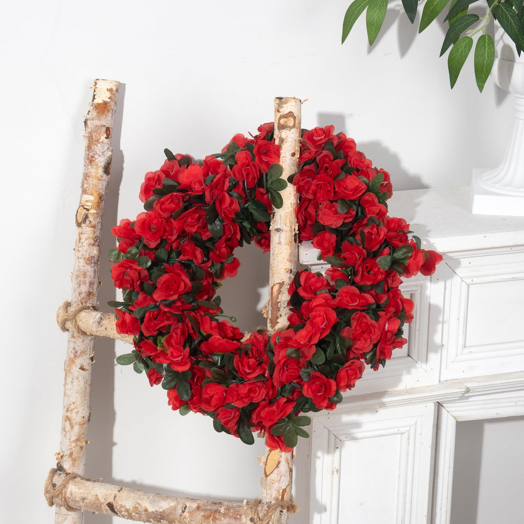 Artificial red rose garland with green leaves draped over a rustic wooden ladder against a white wall.
