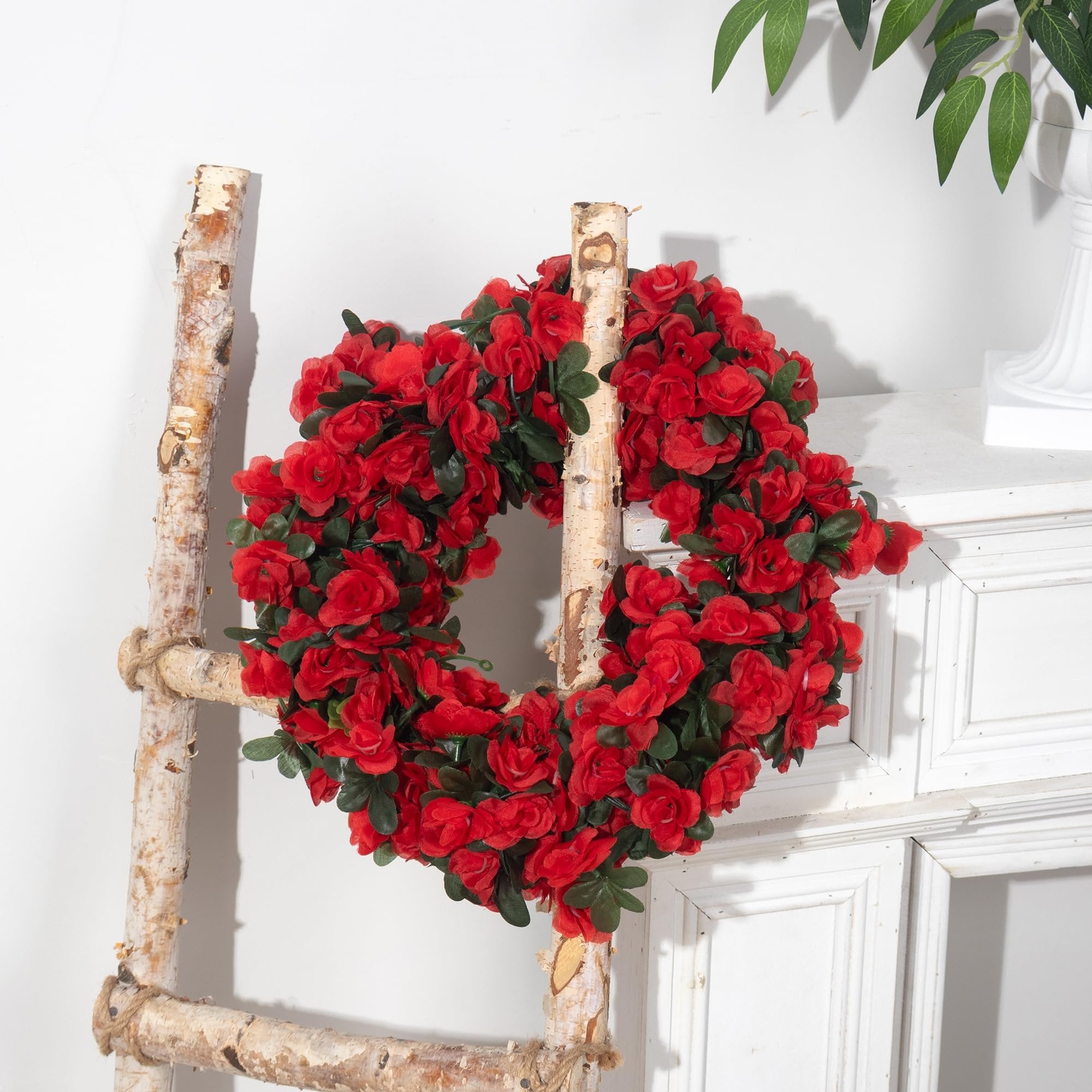 Artificial red rose garland with green leaves draped over a rustic wooden ladder against a white wall.