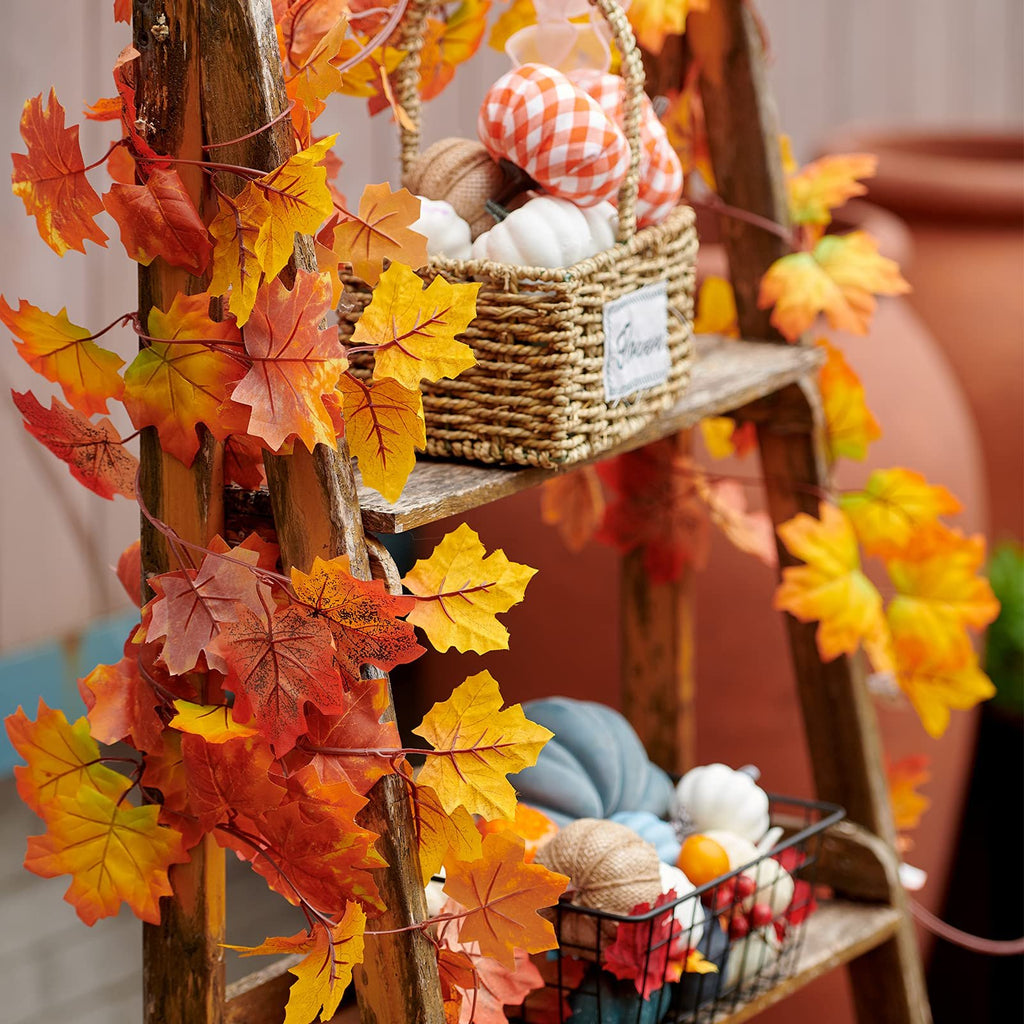 Artificial maple leaf garland draped on a wooden ladder with autumn decor, including pumpkins and gourds in baskets.