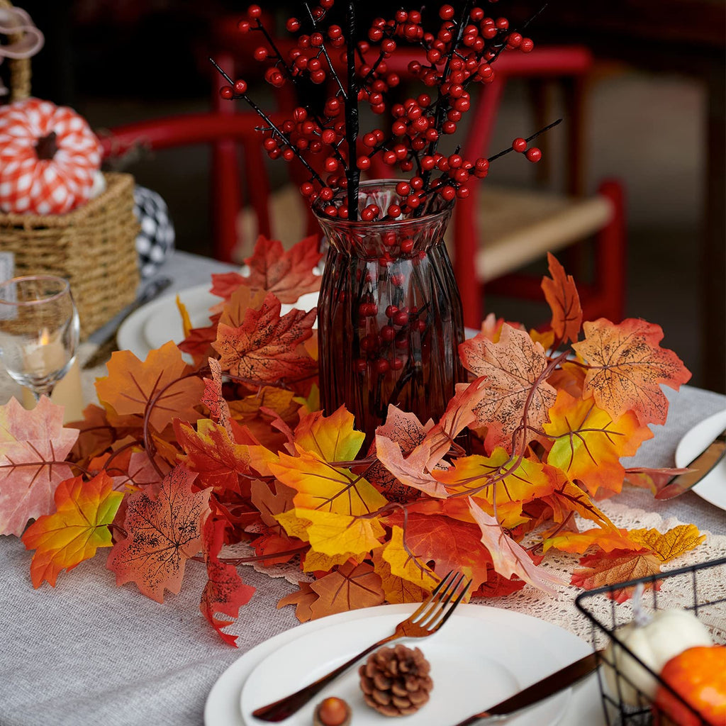 Artificial maple leaf garland in autumn colors arranged on a table with a vase of red berries, plates, and cutlery.