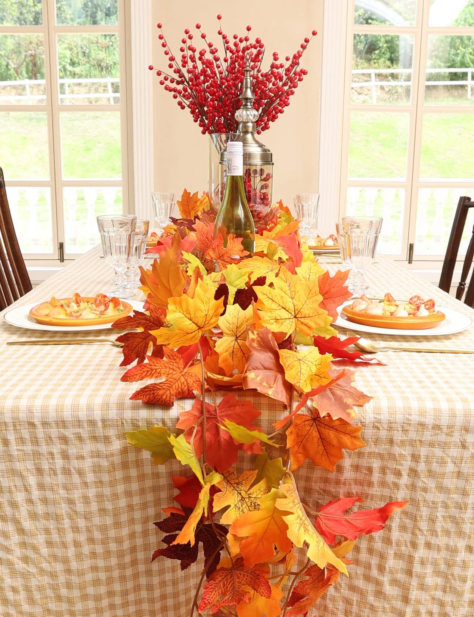 Autumn maple leaf garland draped across a table set for a meal, with glasses, plates, and a centerpiece of red berries.
