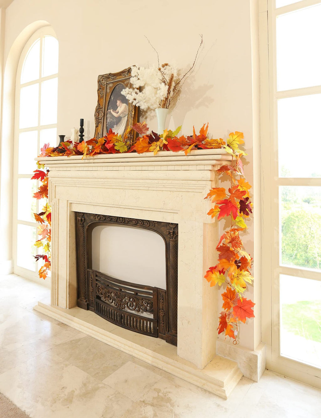 Artificial maple leaf garland draped over a fireplace mantel with decorative items and large windows in the background.