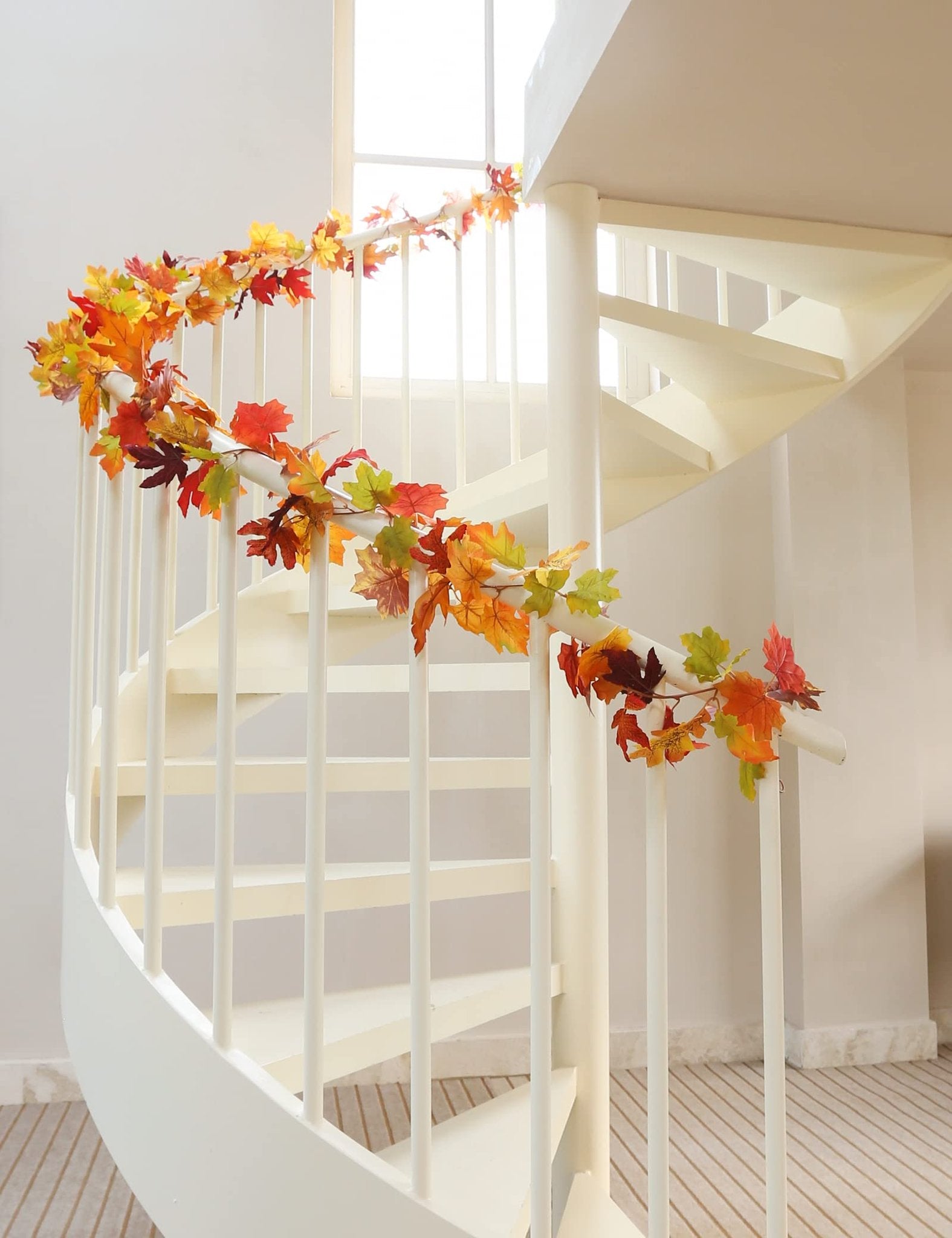 Artificial fall maple leaf garland draped along a white spiral staircase railing, featuring orange, yellow, and red leaves.