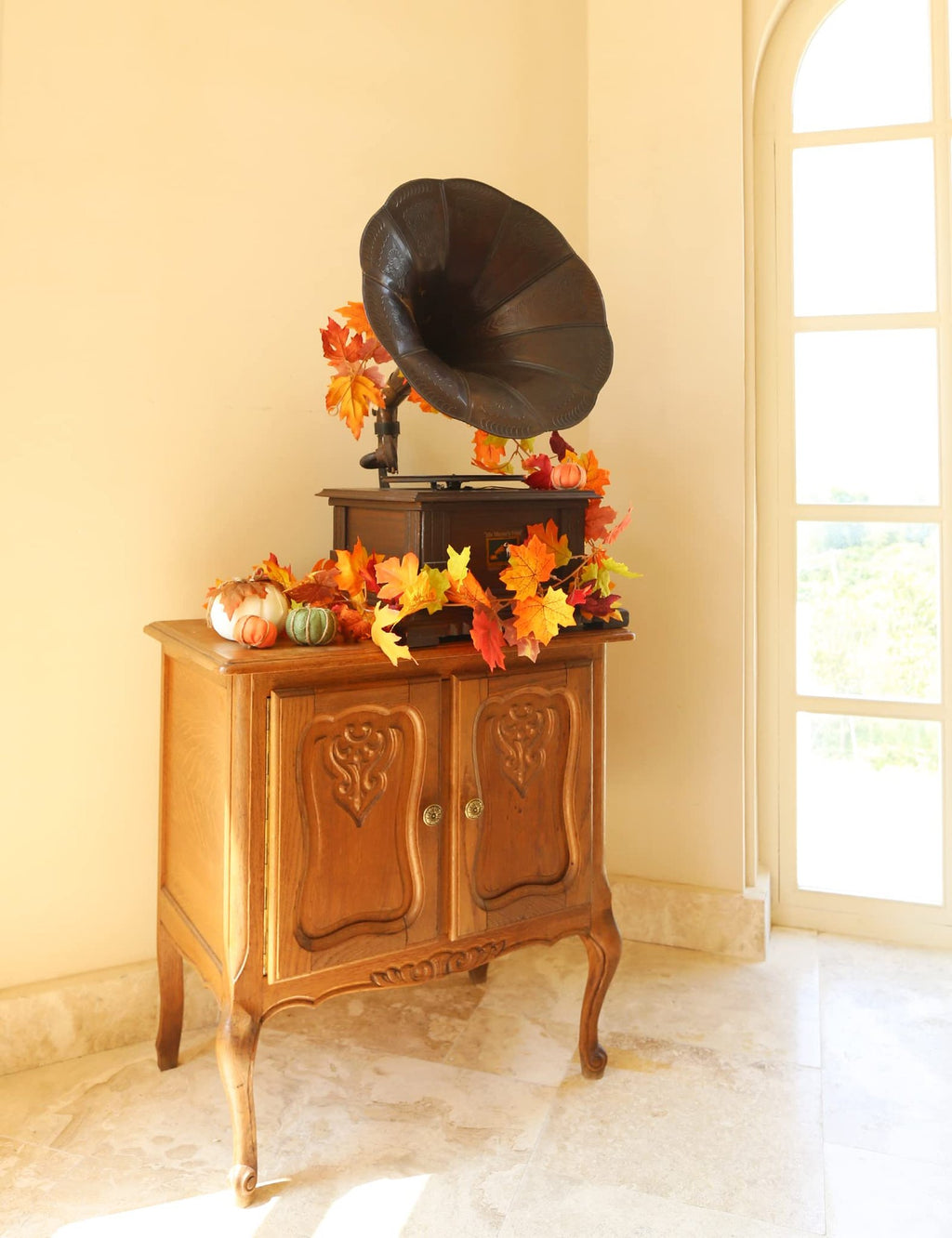 Artificial fall maple leaf garland draped over an antique record player on a wooden cabinet, with decorative pumpkins.