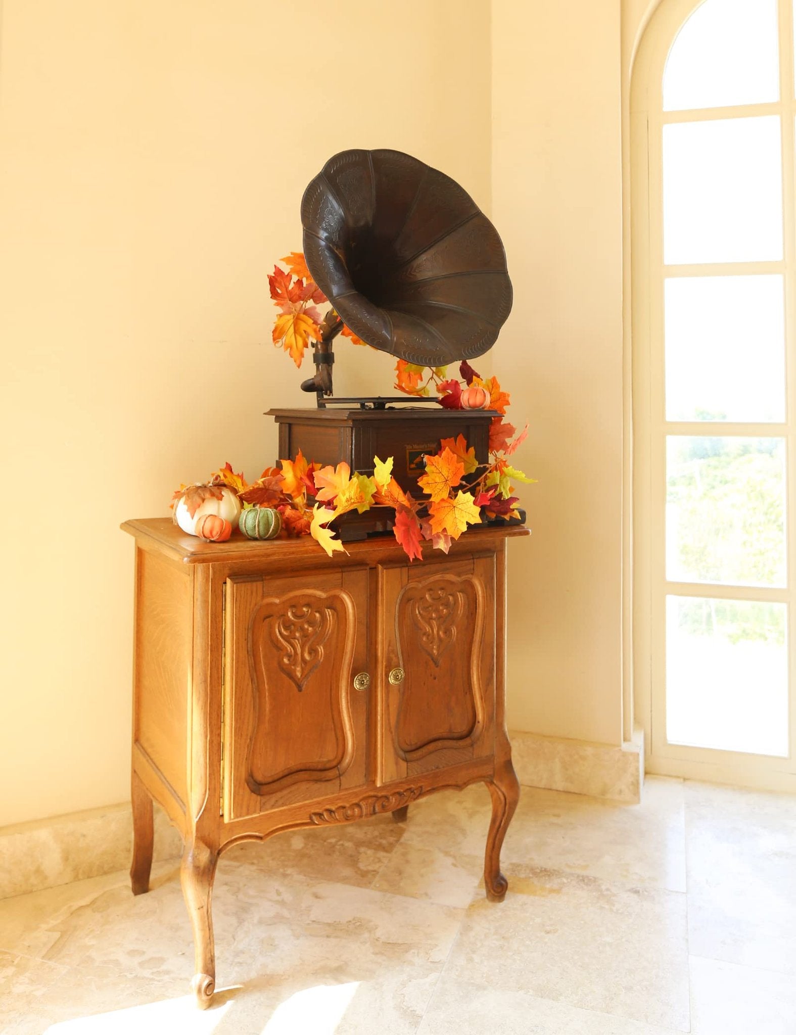 Artificial fall maple leaf garland draped over an antique record player on a wooden cabinet, with decorative pumpkins.