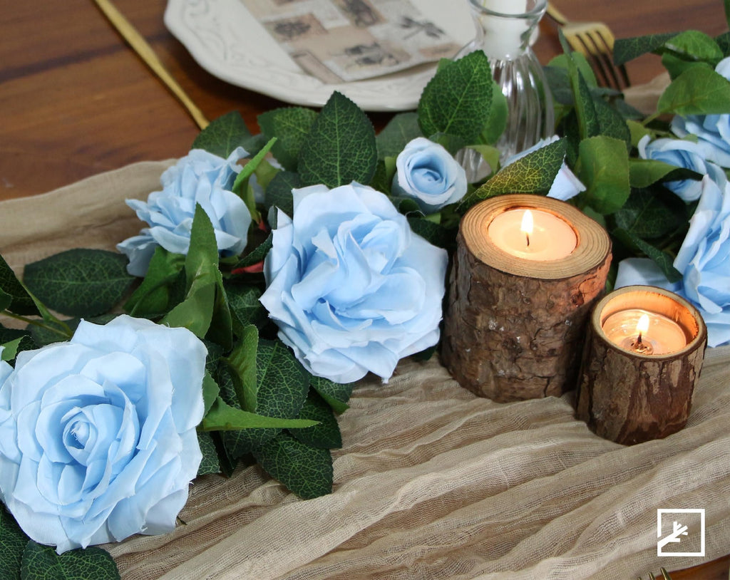 Artificial blue rose garland with green leaves and lit wooden candle holders on a beige runner