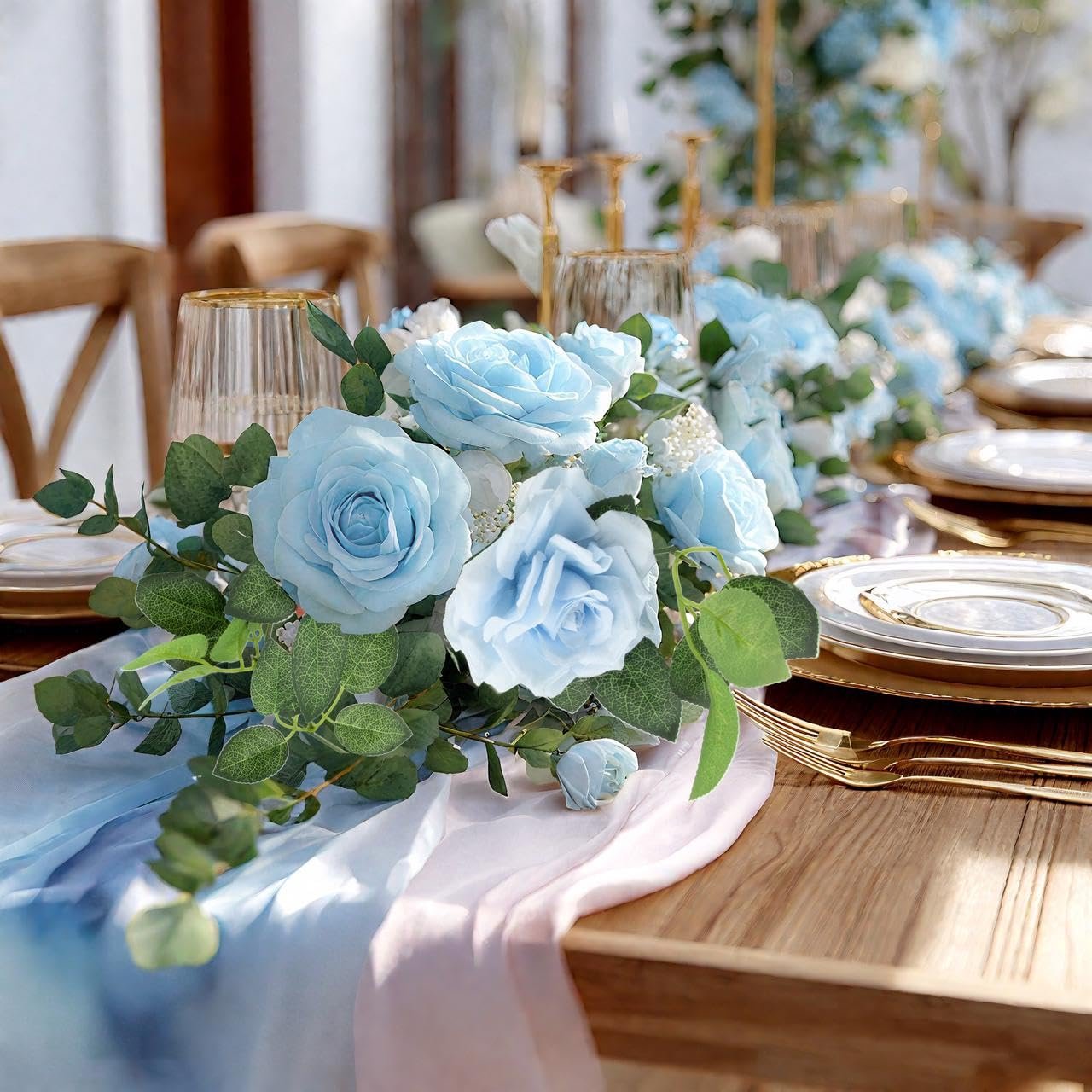 Artificial blue rose garland draped across a wooden table set for a formal dinner, with gold-rimmed plates and cutlery.