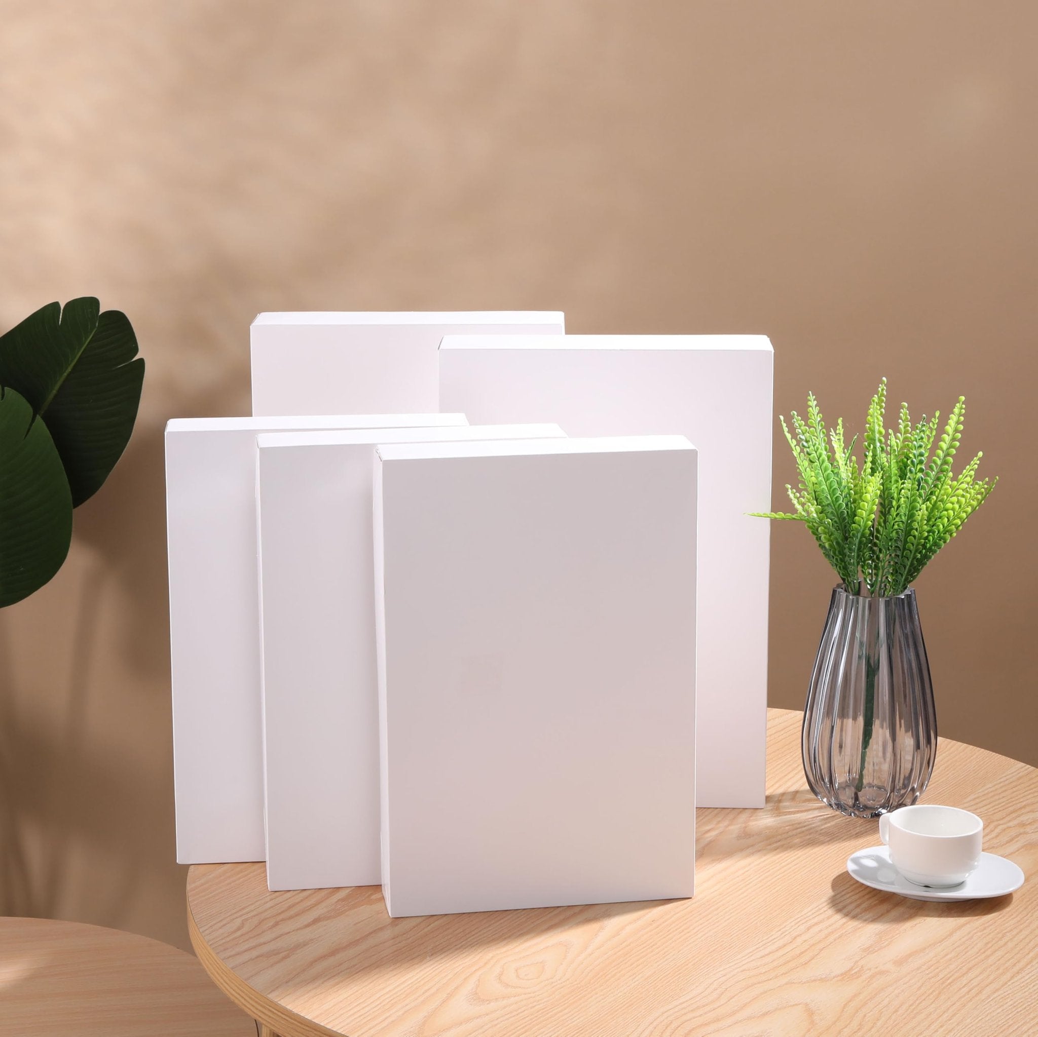 Five white gift boxes of varying sizes arranged on a wooden table with a plant and a teacup.