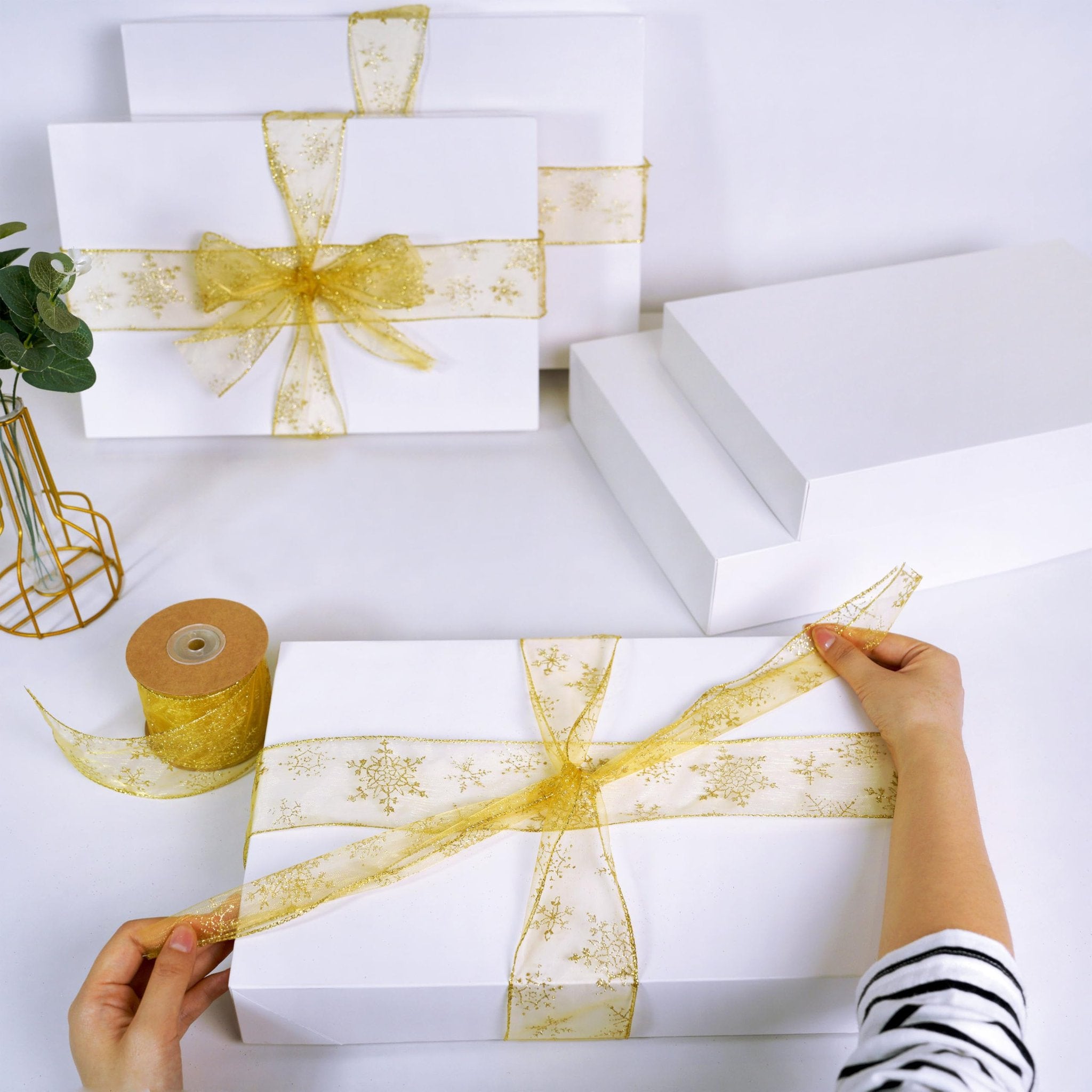 Hands tying gold snowflake ribbon around white gift boxes of assorted sizes on a white surface