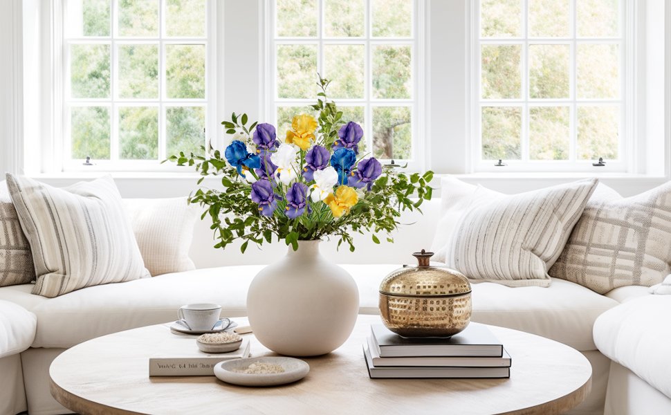 Bouquet of artificial iris flowers in a vase on a coffee table with pillows and books.