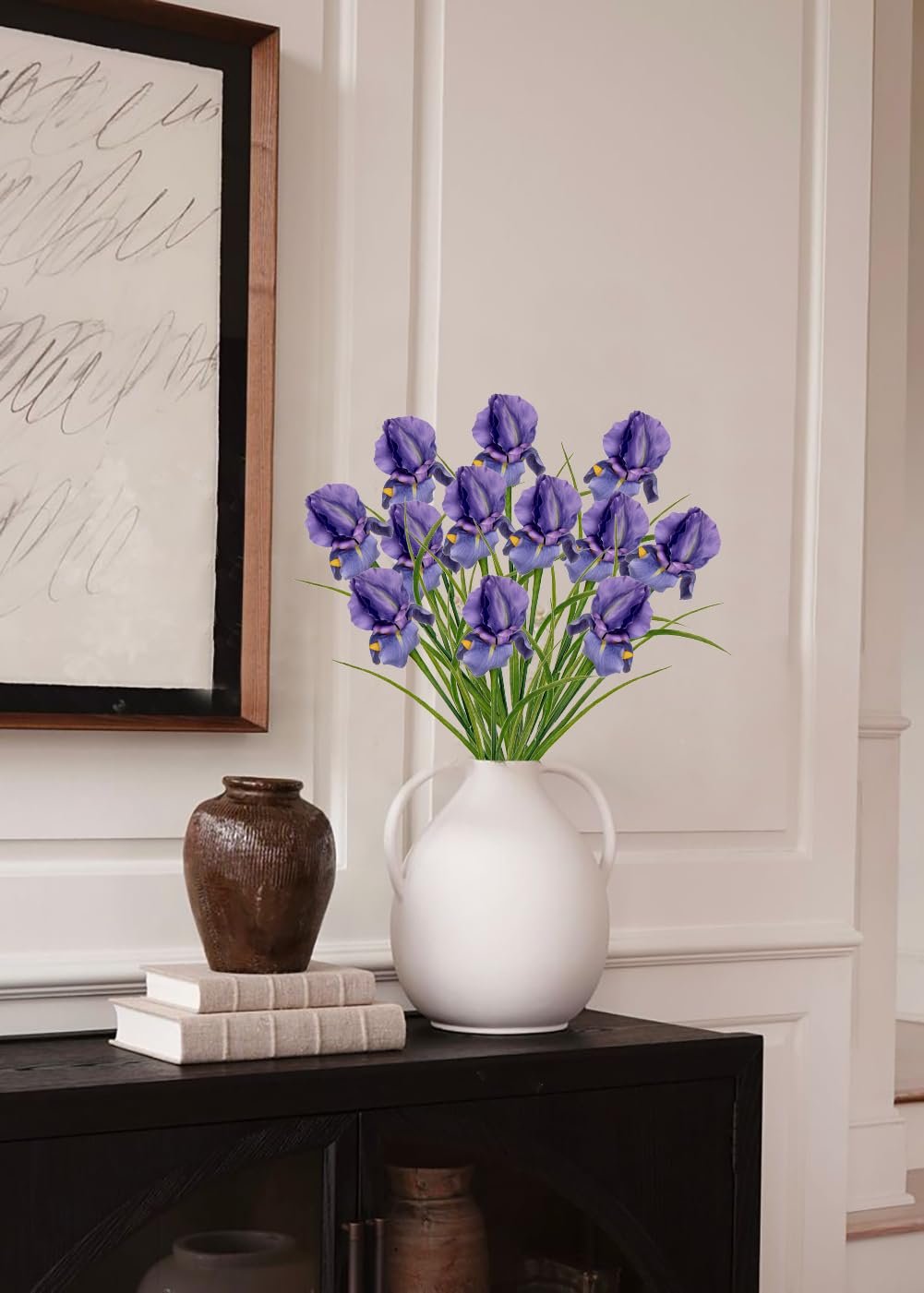 A bouquet of purple artificial iris flowers in a white vase on a dark cabinet, next to books and a decorative jar.