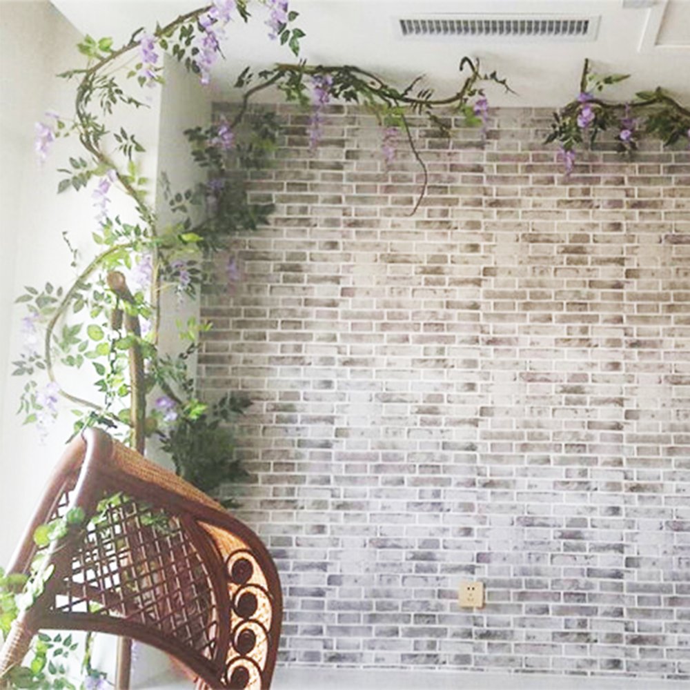 Artificial purple wisteria garland draped along a wall and over a wicker chair, with a brick pattern background.