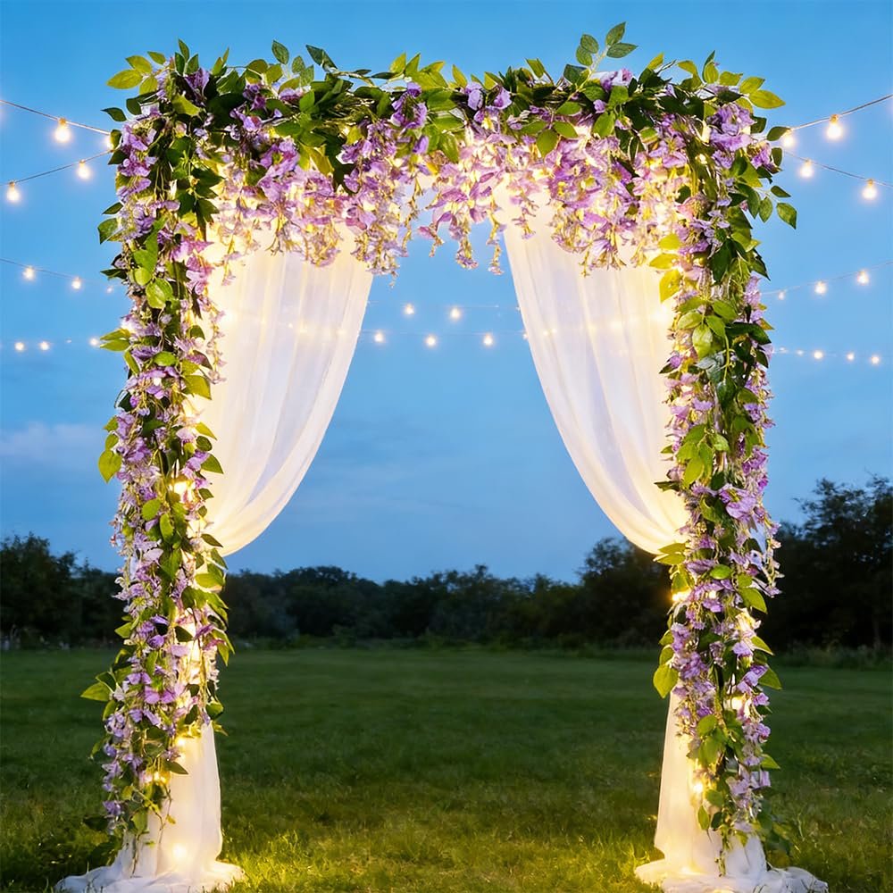 Wedding arch decorated with purple wisteria garlands, white sheer fabric, and string lights at dusk