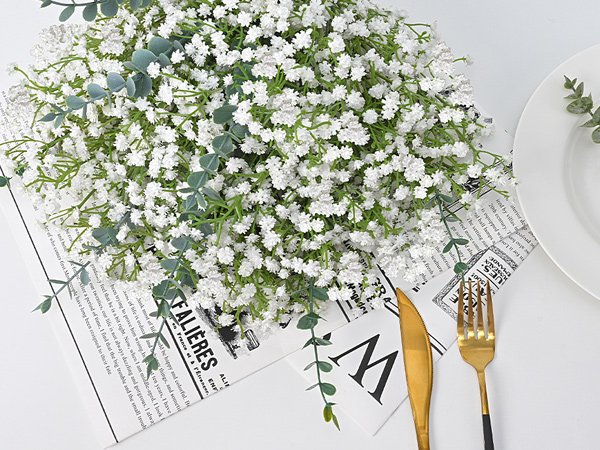 Close-up of white artificial baby's breath flowers with eucalyptus sprigs, arranged on a surface with newspaper and cutlery.