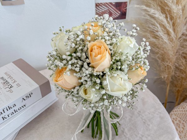 Artificial baby's breath and rose bouquet with white and peach flowers tied with ribbon, next to books and dried reeds.