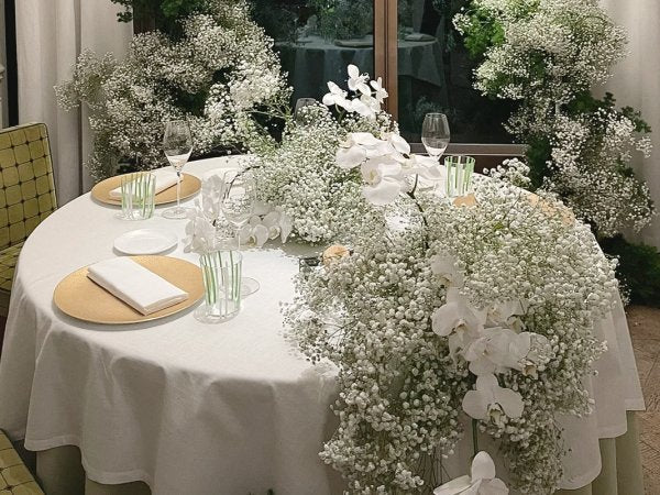 Round table set for a wedding reception with white tablecloth, gold plates, white napkins, and abundant white baby's breath and orchid floral arrangements.