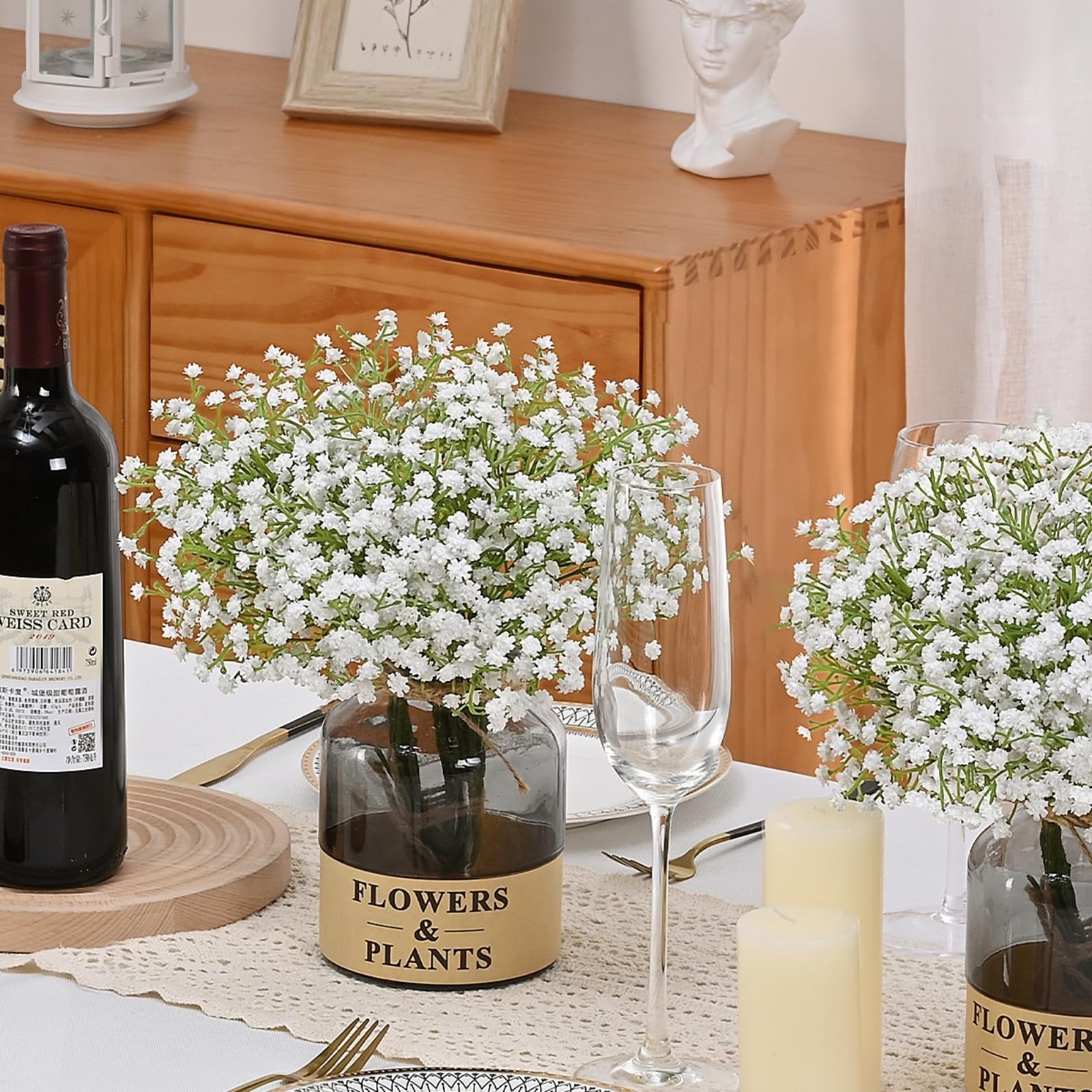 Two vases of white artificial baby's breath flowers on a table setting with a bottle of red wine and champagne glasses.