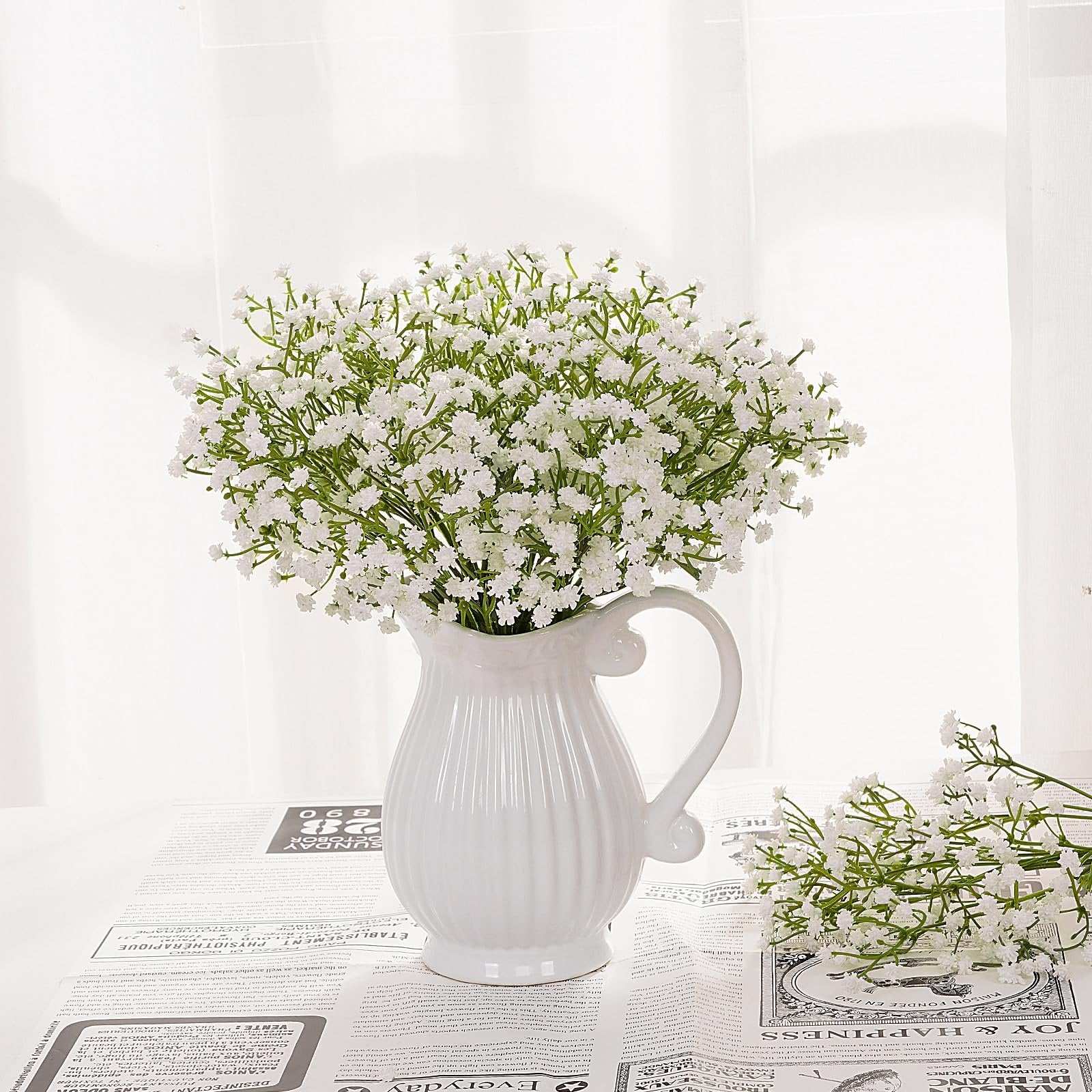 White artificial baby's breath flowers in a white pitcher on a newspaper-like surface.