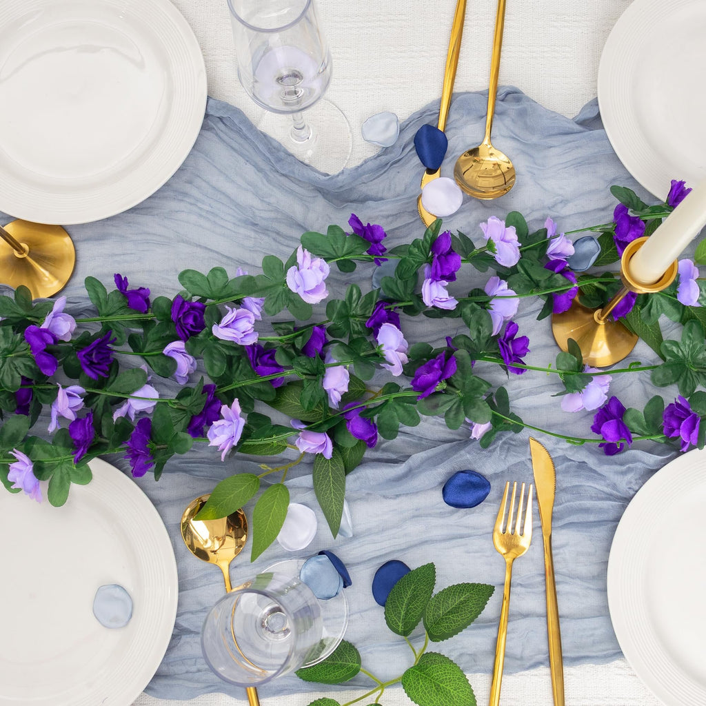 Artificial purple rose garland draped over a table setting with plates, gold cutlery, and wine glasses.