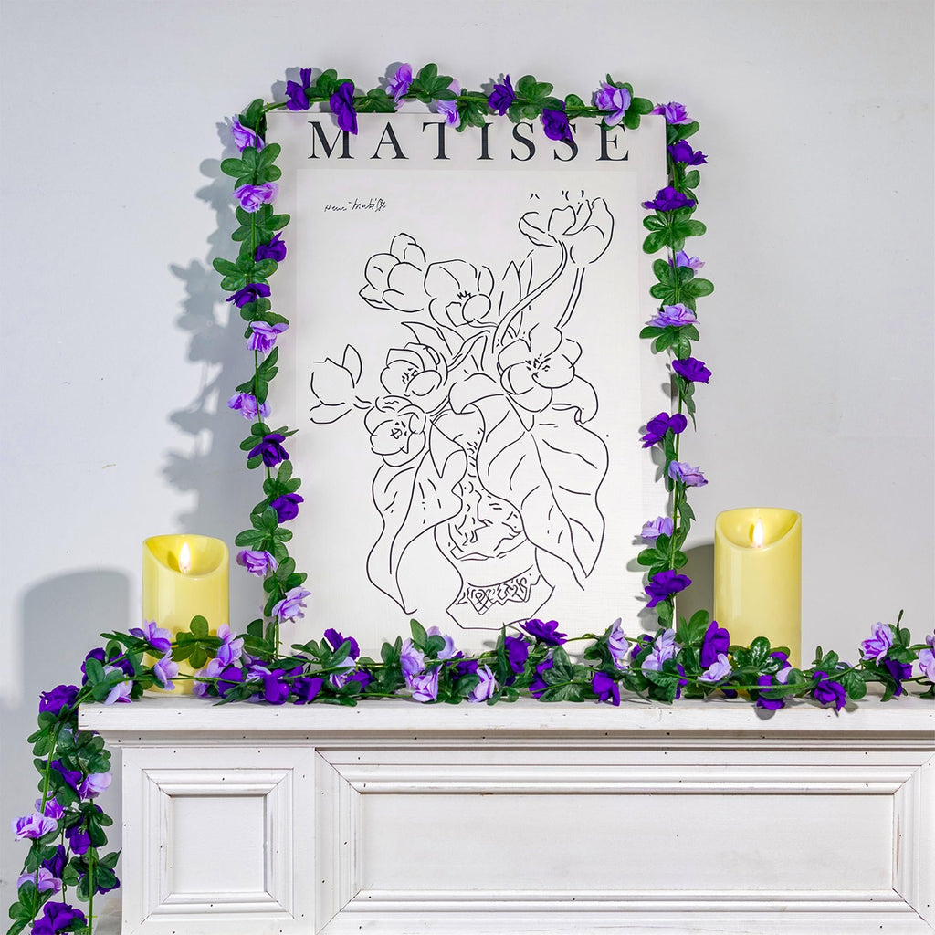 Artificial purple rose garland draped around a framed Matisse print and two pillar candles on a mantelpiece.