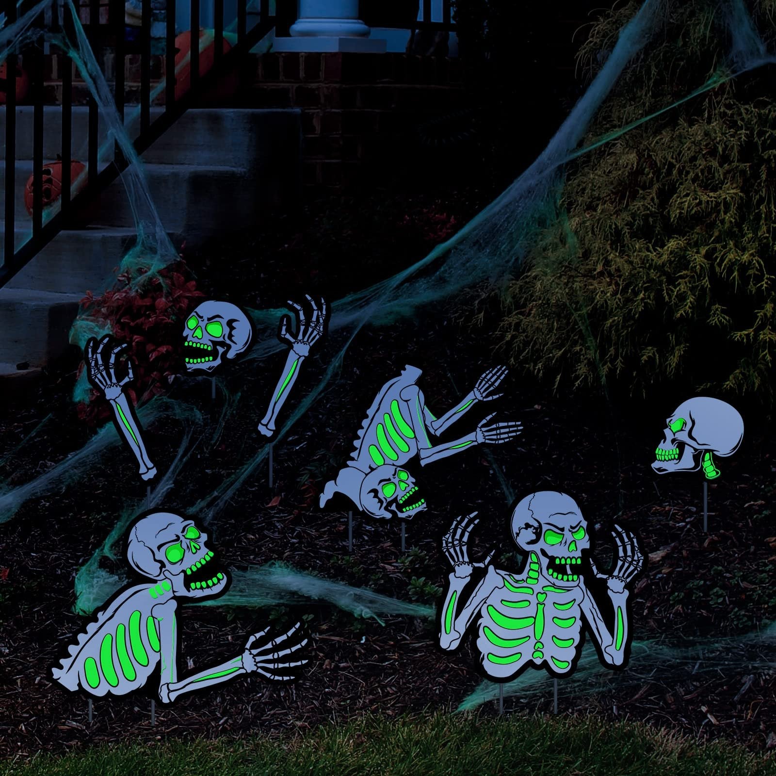 Fluorescent skeleton yard decorations with green glowing eyes and ribs, arranged on a lawn with cobwebs and near stairs.