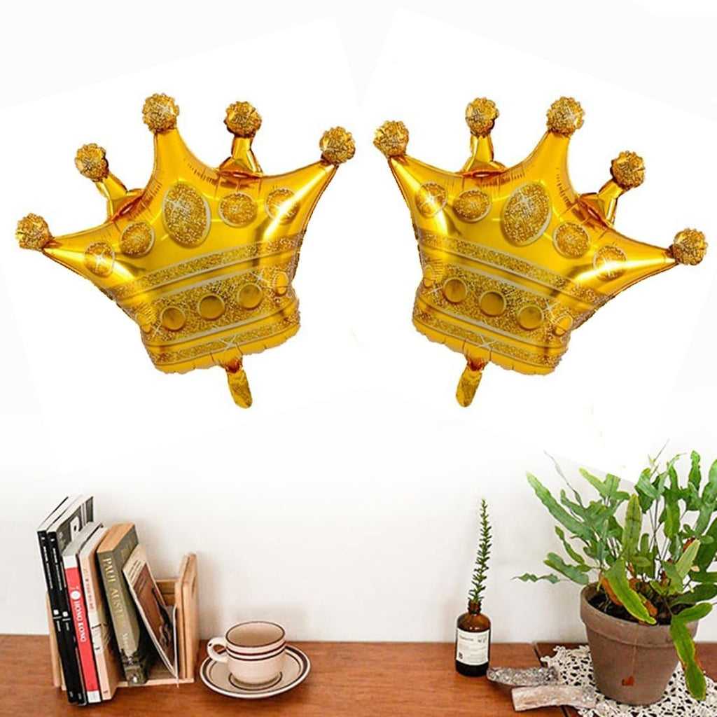 Two gold foil crown-shaped balloons with glitter accents, displayed on a white background above a shelf with books and a plant.