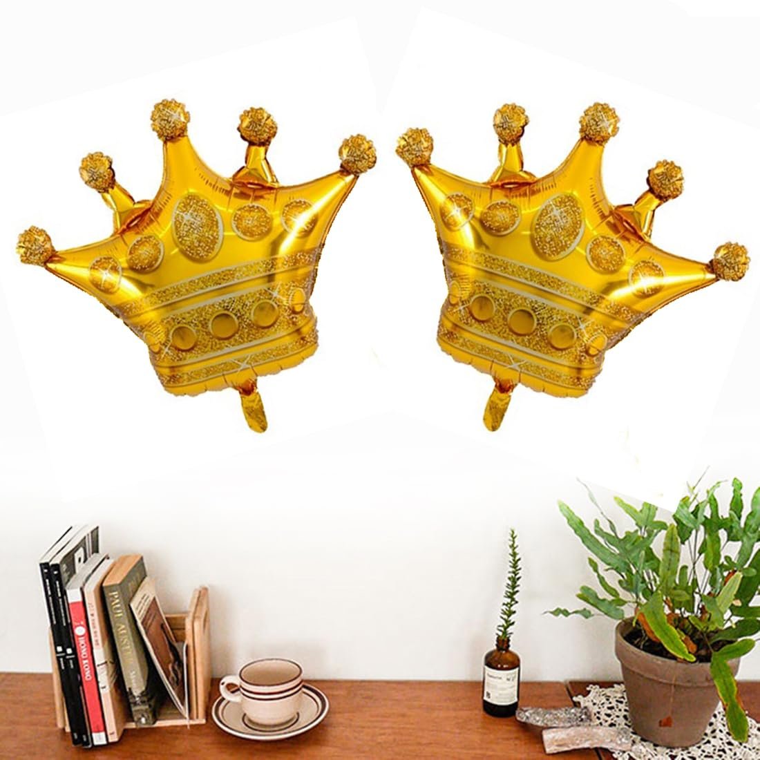Two gold foil crown-shaped balloons with glitter accents, displayed on a white background above a shelf with books and a plant.