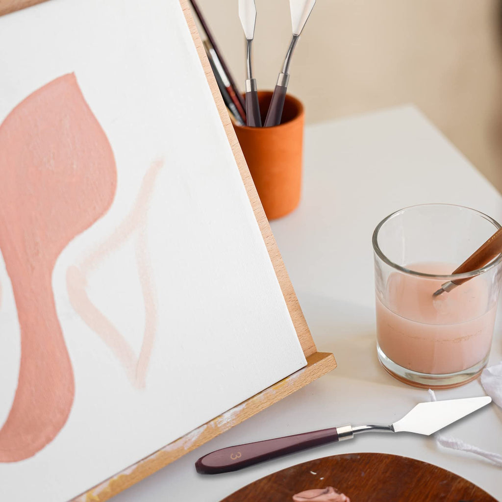 Artist's easel with a painting in progress, palette knives in a cup, and a glass of liquid on a white table.