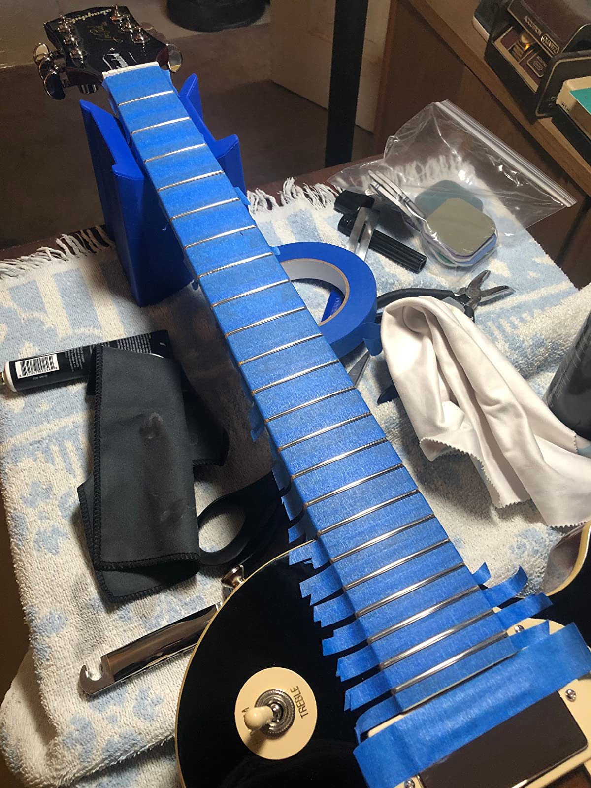 Guitar neck with frets taped off with blue masking tape, ready for fret polishing. Tools and cloth are nearby.
