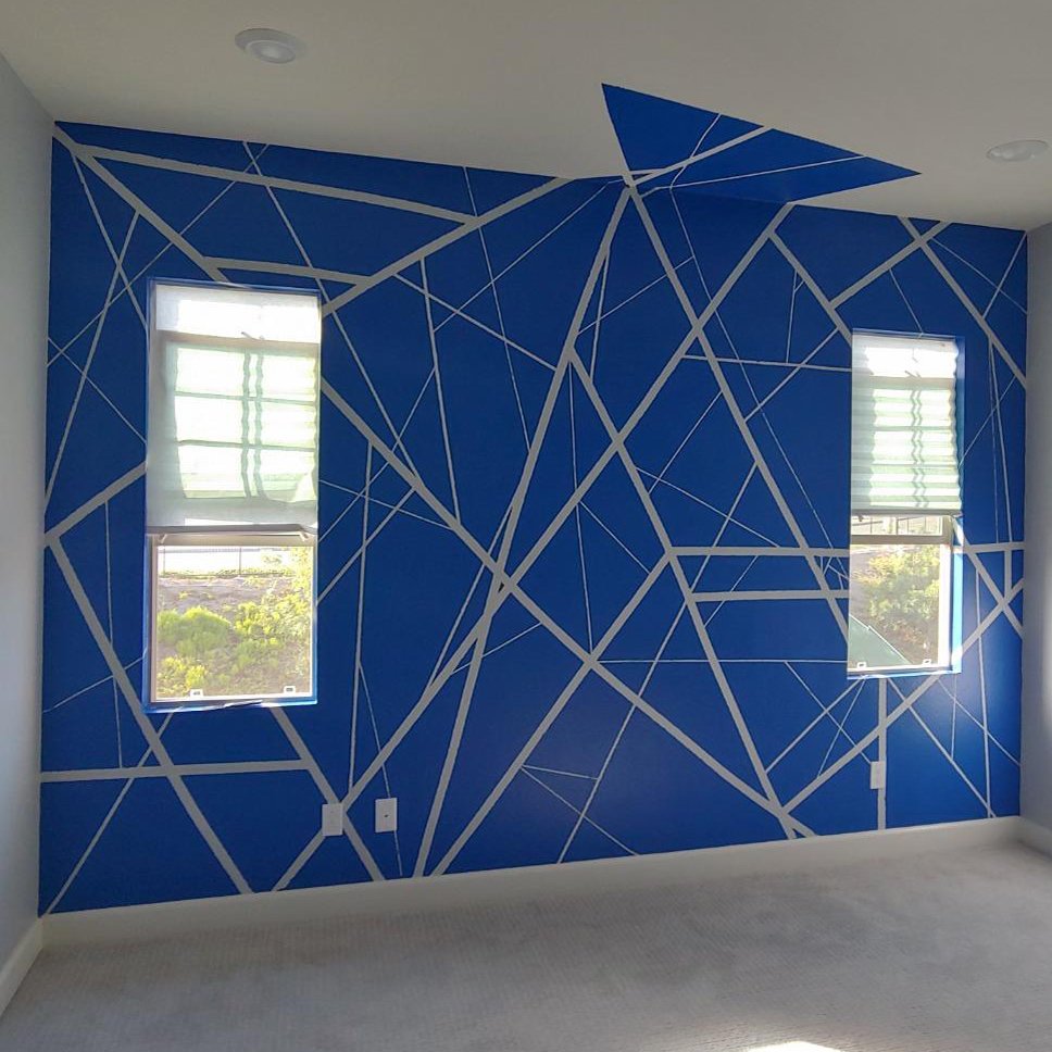 Room with blue walls featuring a geometric pattern created with white masking tape, two windows with blinds, and carpeted floor.