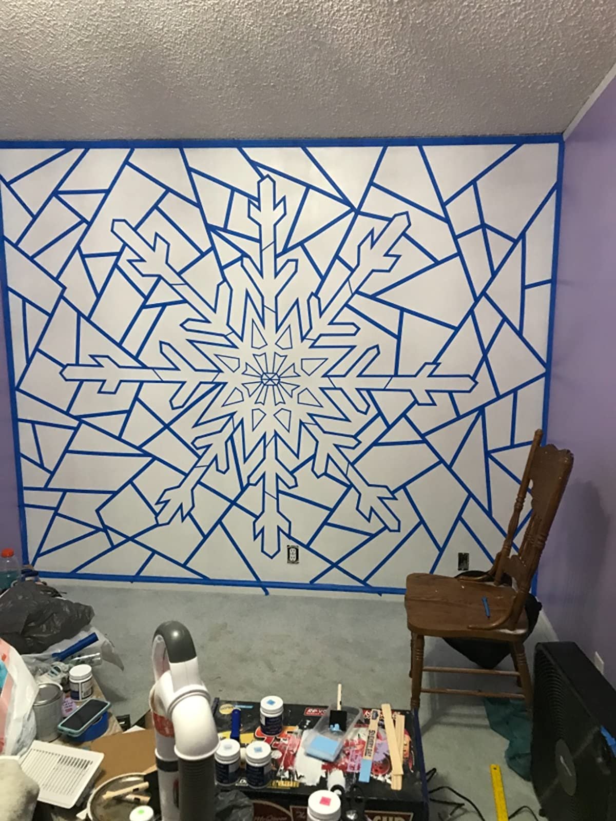 Wall mural design using blue tape to create a snowflake pattern on a white wall, with a chair and art supplies in the foreground.