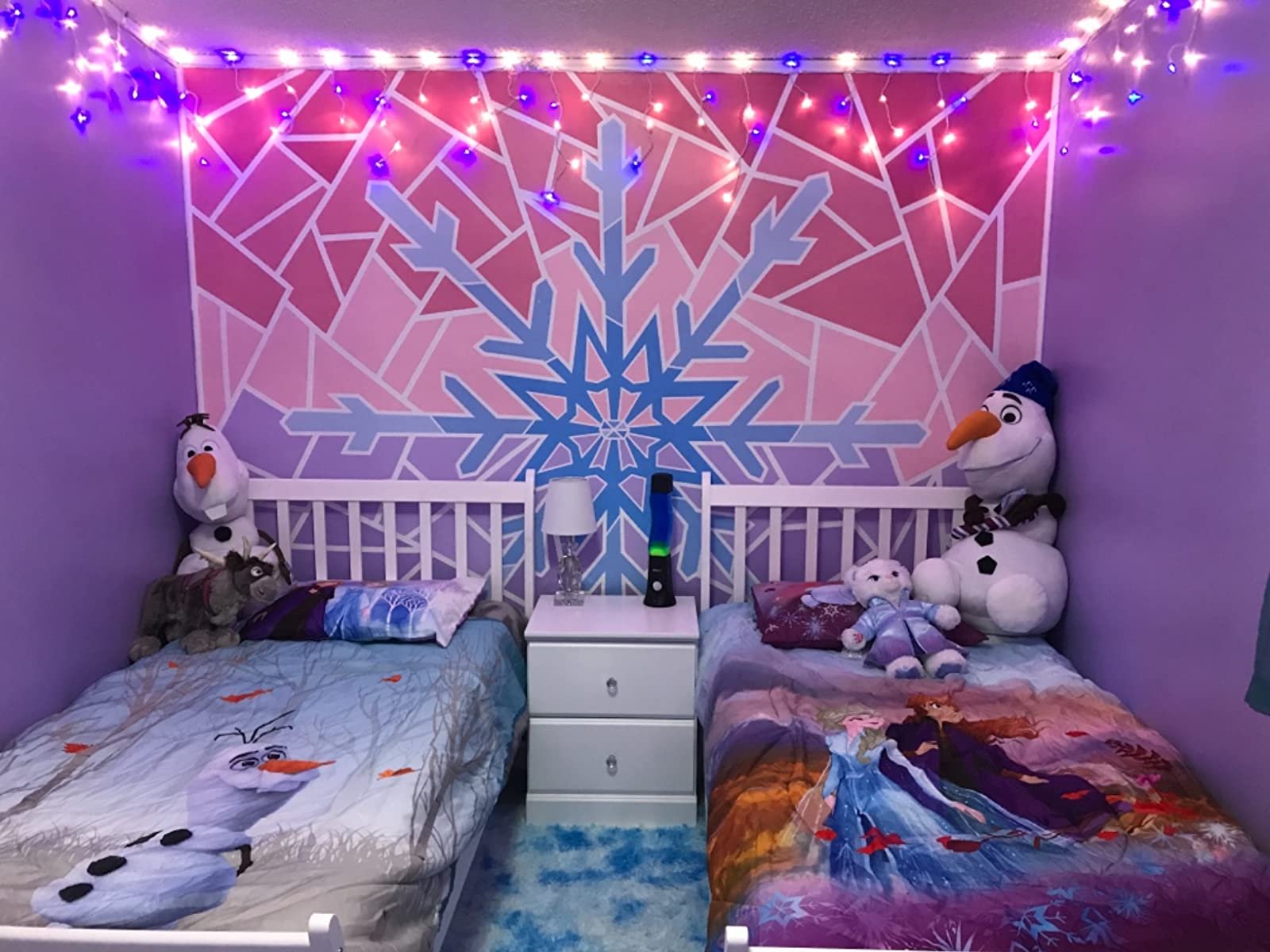 Two beds in a room decorated with a large snowflake mural and string lights, featuring Frozen characters on bedding and plush toys.