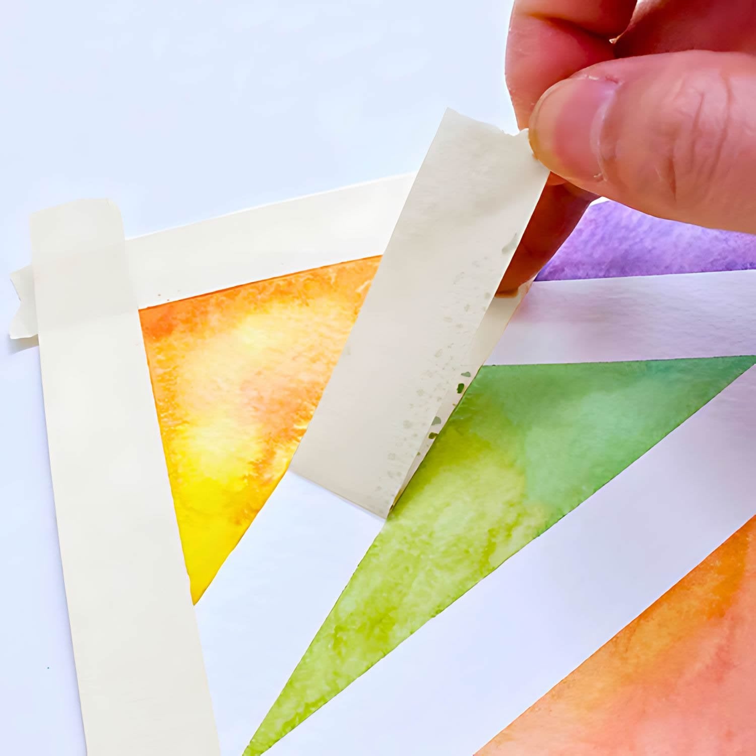 Hand peeling masking tape from a watercolor painting with orange, yellow, green, and purple washes.