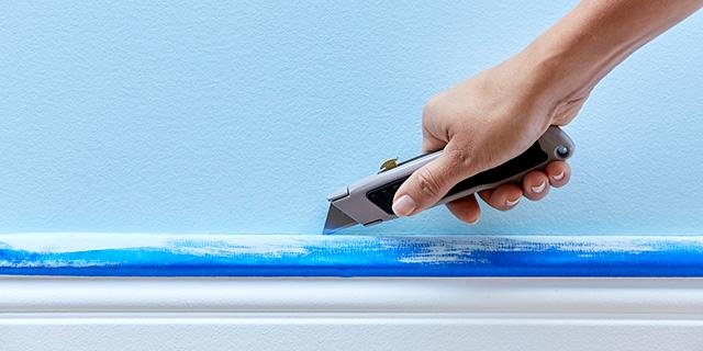 Hand using a utility knife to cut blue painter's tape along a white baseboard against a light blue wall.