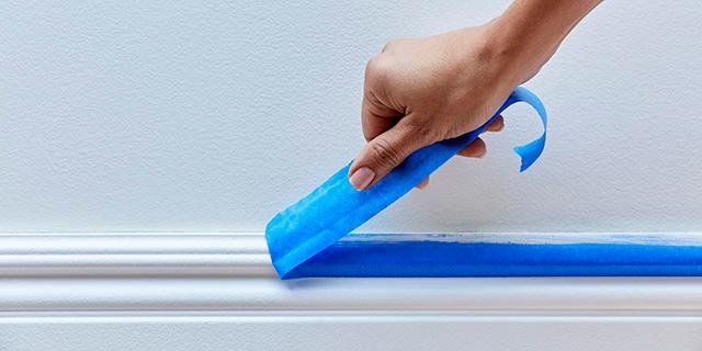 Hand applying blue painter's tape along a white baseboard for painting.