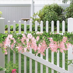 Pink wisteria garland with green leaves draped over a white picket fence in a garden setting.