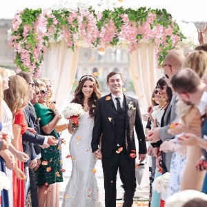 Wedding couple walking through an arch decorated with pink wisteria garlands, showered with petals by guests.