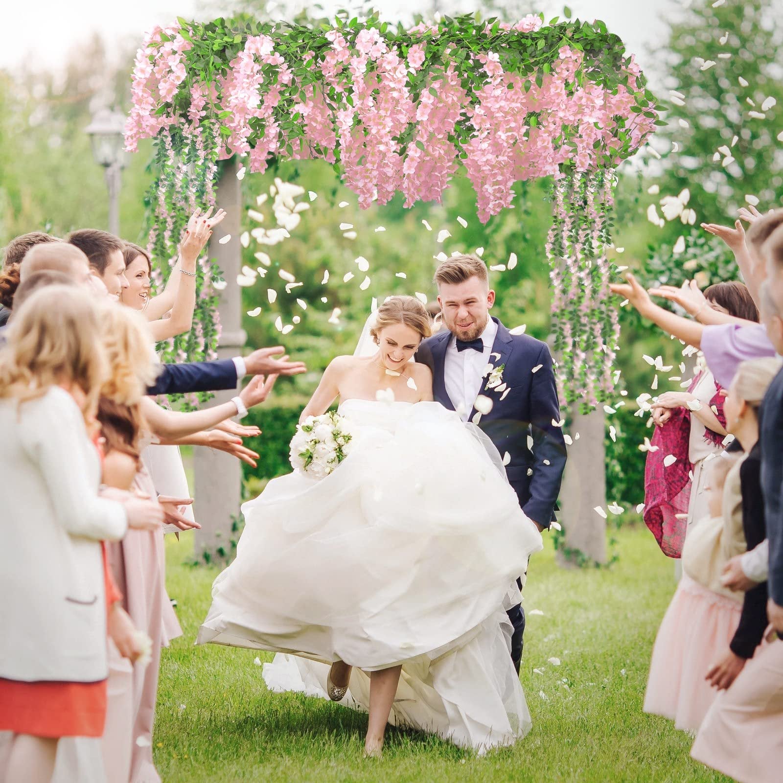 Newlyweds walking through a petal shower under a pink wisteria floral archway at an outdoor wedding ceremony.