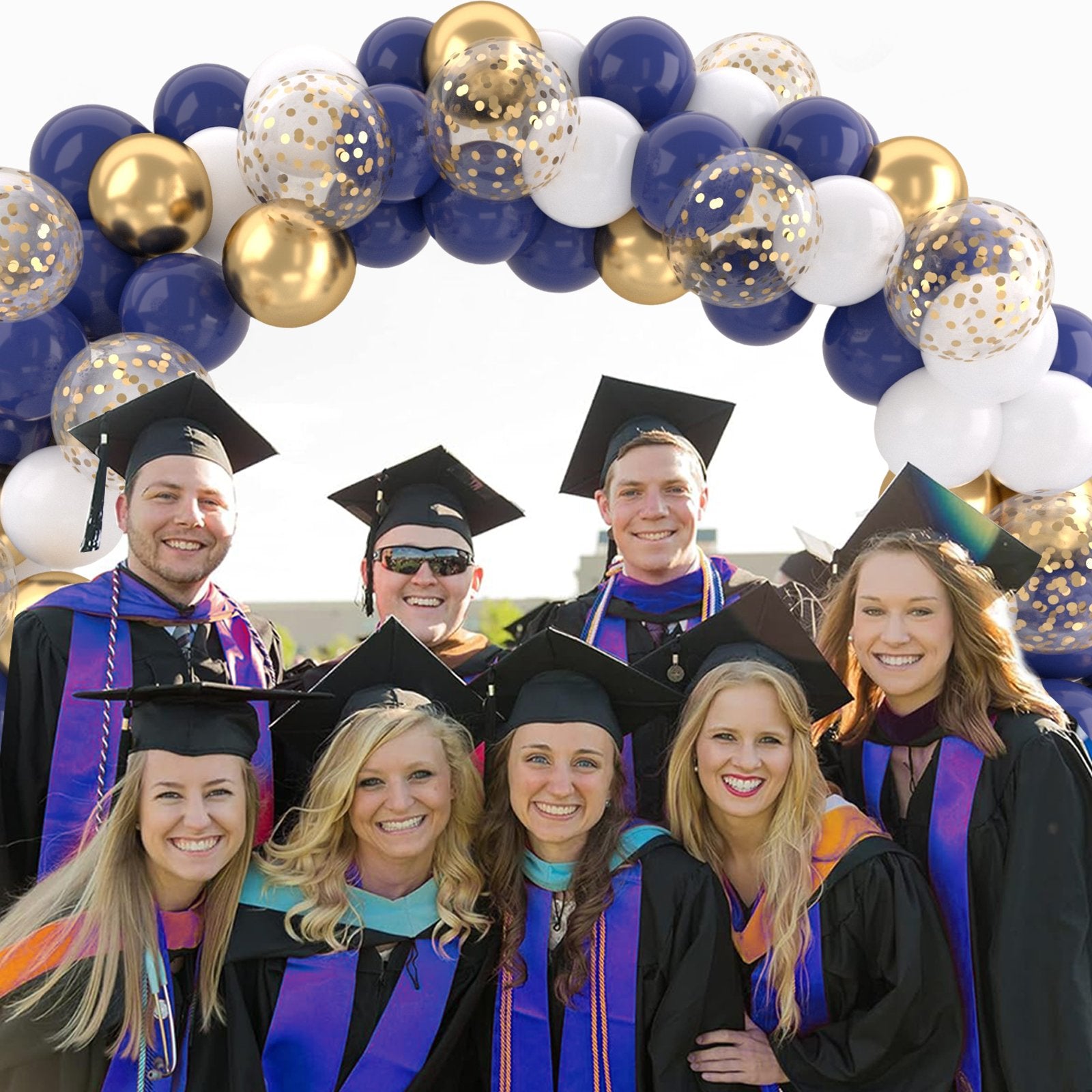 Graduating students in caps and gowns pose under a balloon arch featuring navy blue, gold, and white confetti balloons.